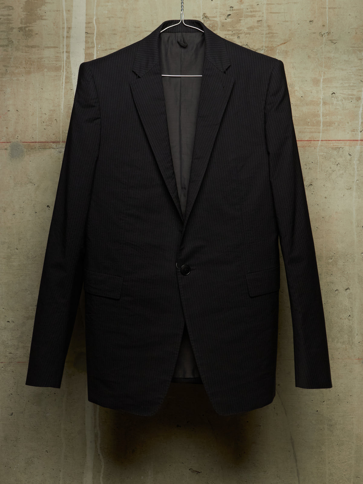 A black pinstripe suit jacket hanging on a metal hanger against a textured concrete wall.