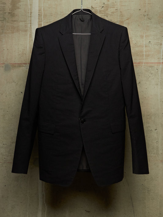 A black pinstripe suit jacket hanging on a metal hanger against a textured concrete wall.
