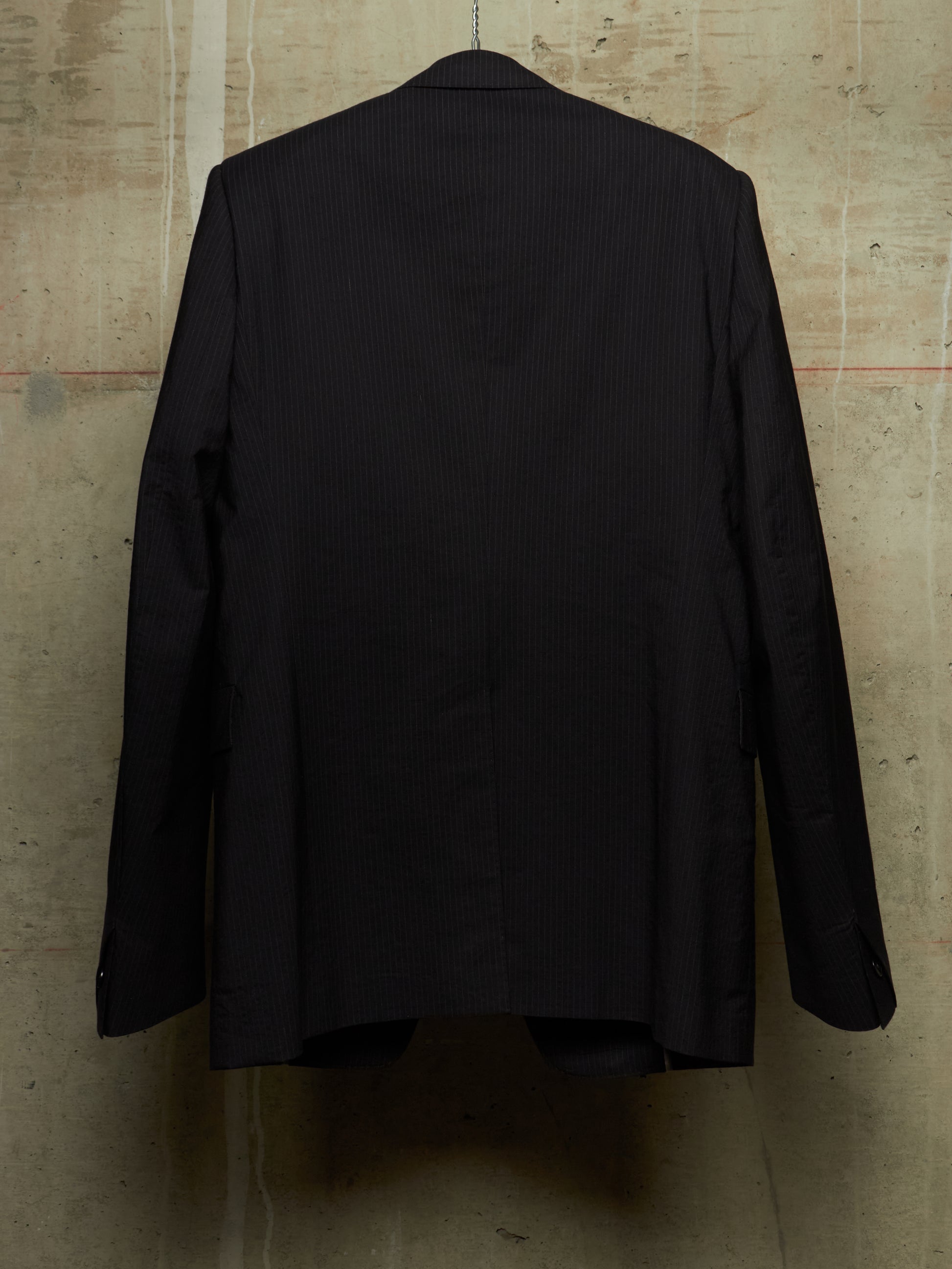 A black pinstripe suit jacket hanging on a hanger against a textured concrete wall, showing the back view.