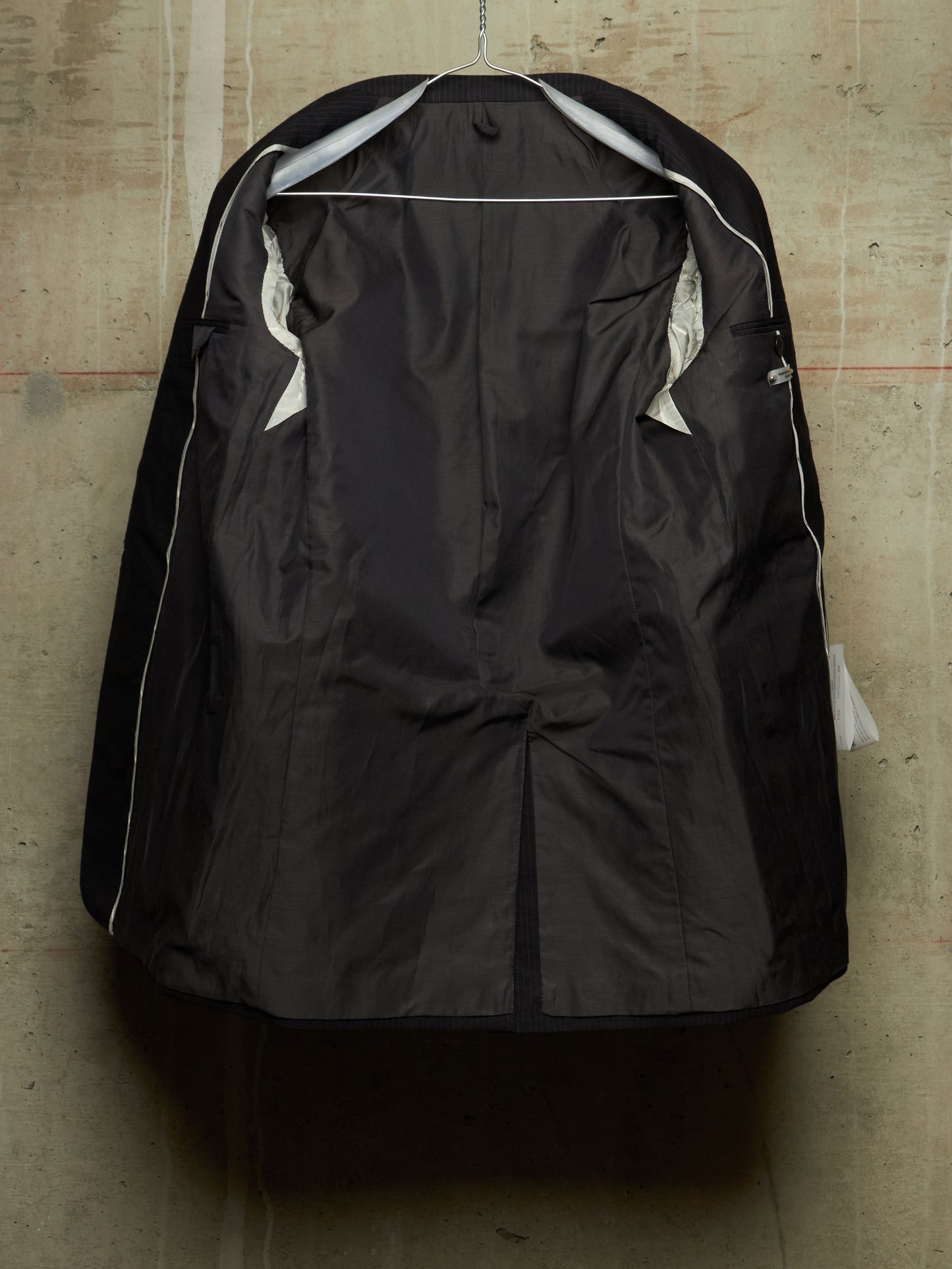 A black waterproof jacket cover hanging on a metal hanger against a textured concrete wall. The cover is shown from the back, featuring a central seam, zippered pockets, and white piping details along the edges.