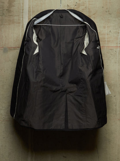 A black waterproof jacket cover hanging on a metal hanger against a textured concrete wall. The cover is shown from the back, featuring a central seam, zippered pockets, and white piping details along the edges.