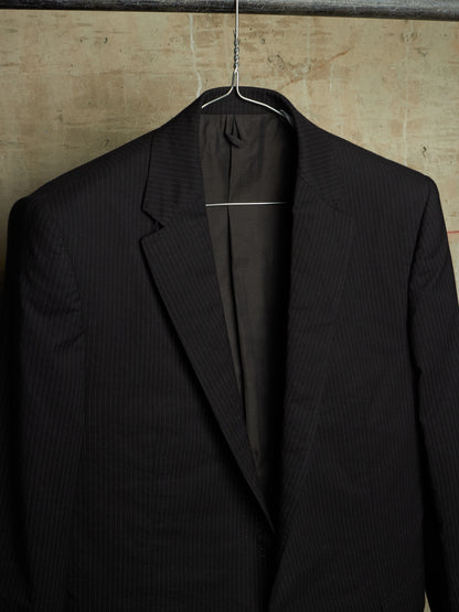 A black pinstripe suit jacket hanging on a wire hanger against a textured beige wall.
