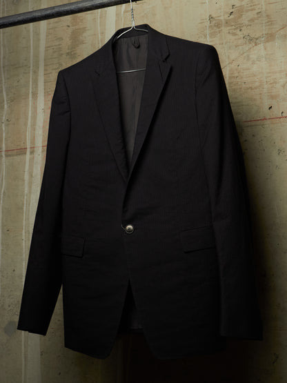 A black single-breasted suit jacket hanging on a hanger against a textured concrete wall.