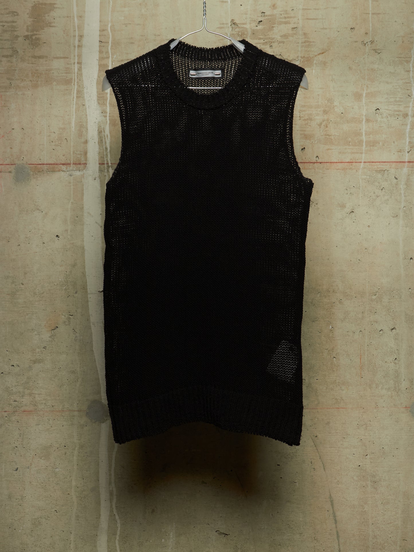 A black sleeveless knit vest with a textured open-knit pattern, hanging on a wire hanger against a concrete wall.