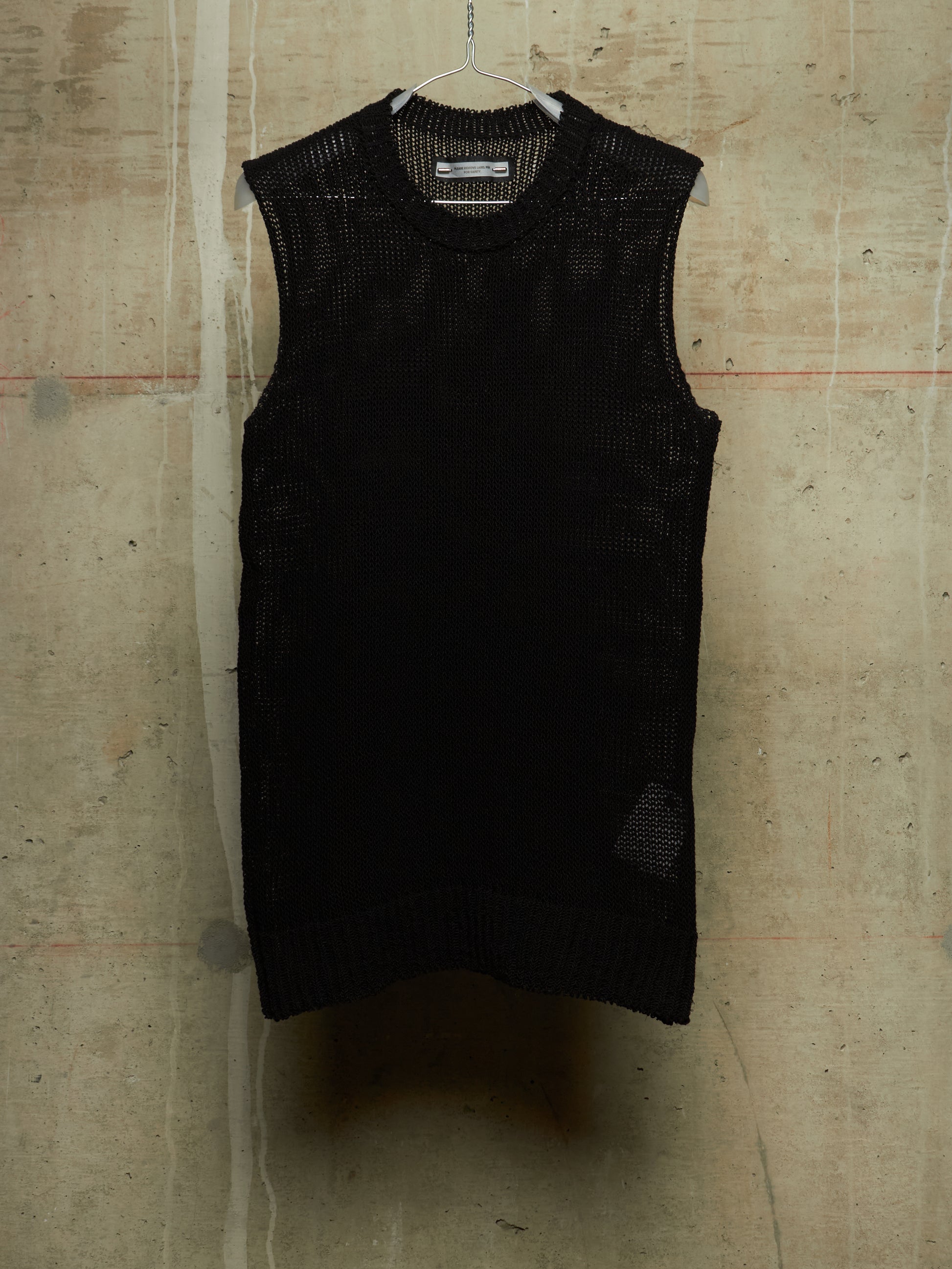 A black sleeveless knit vest with a textured open-knit pattern, hanging on a wire hanger against a concrete wall.