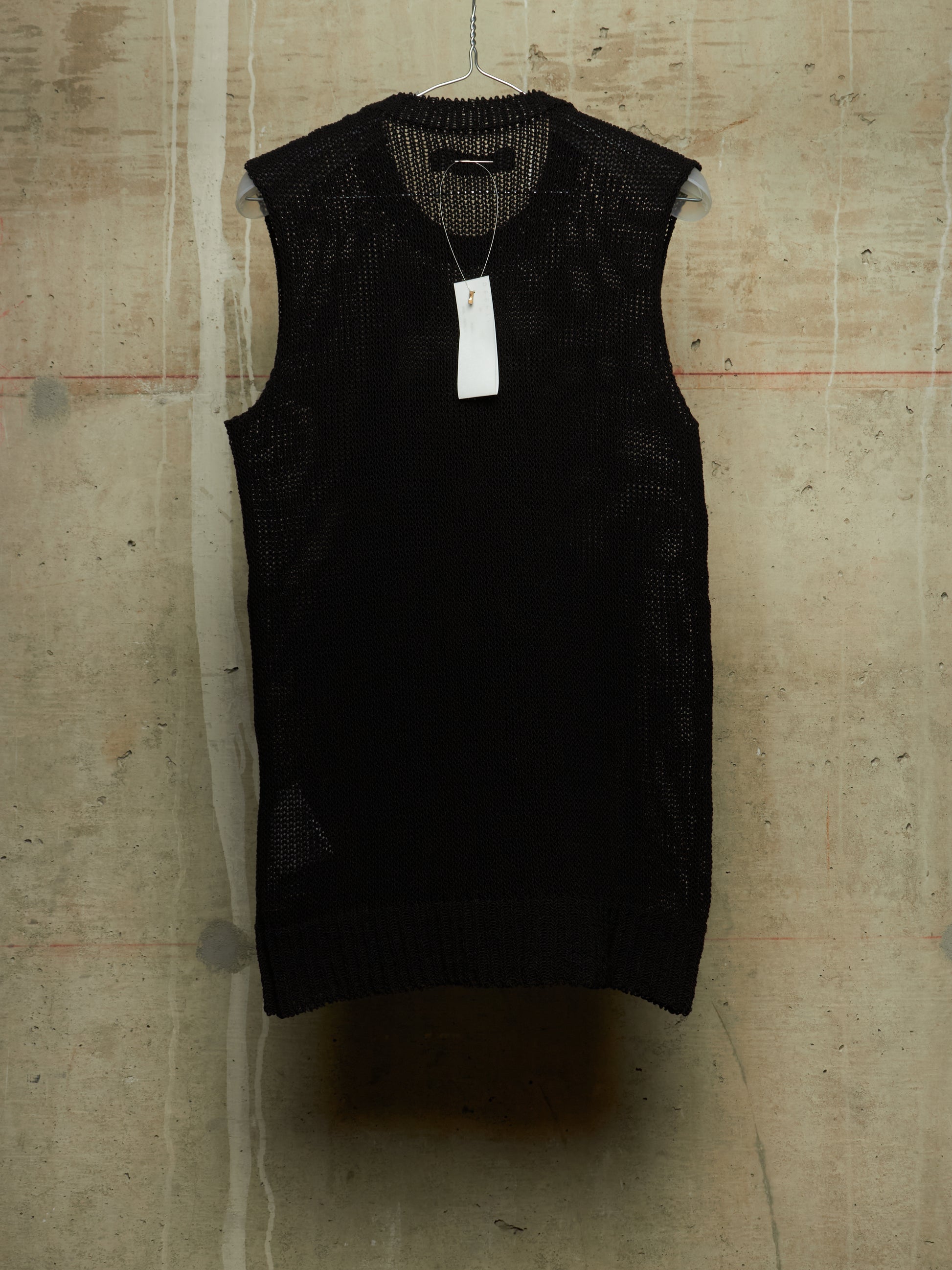 A black sleeveless knit vest with a textured open-knit pattern, hanging on a wire hanger against a concrete wall. A white paper price tag is attached to the neckline.