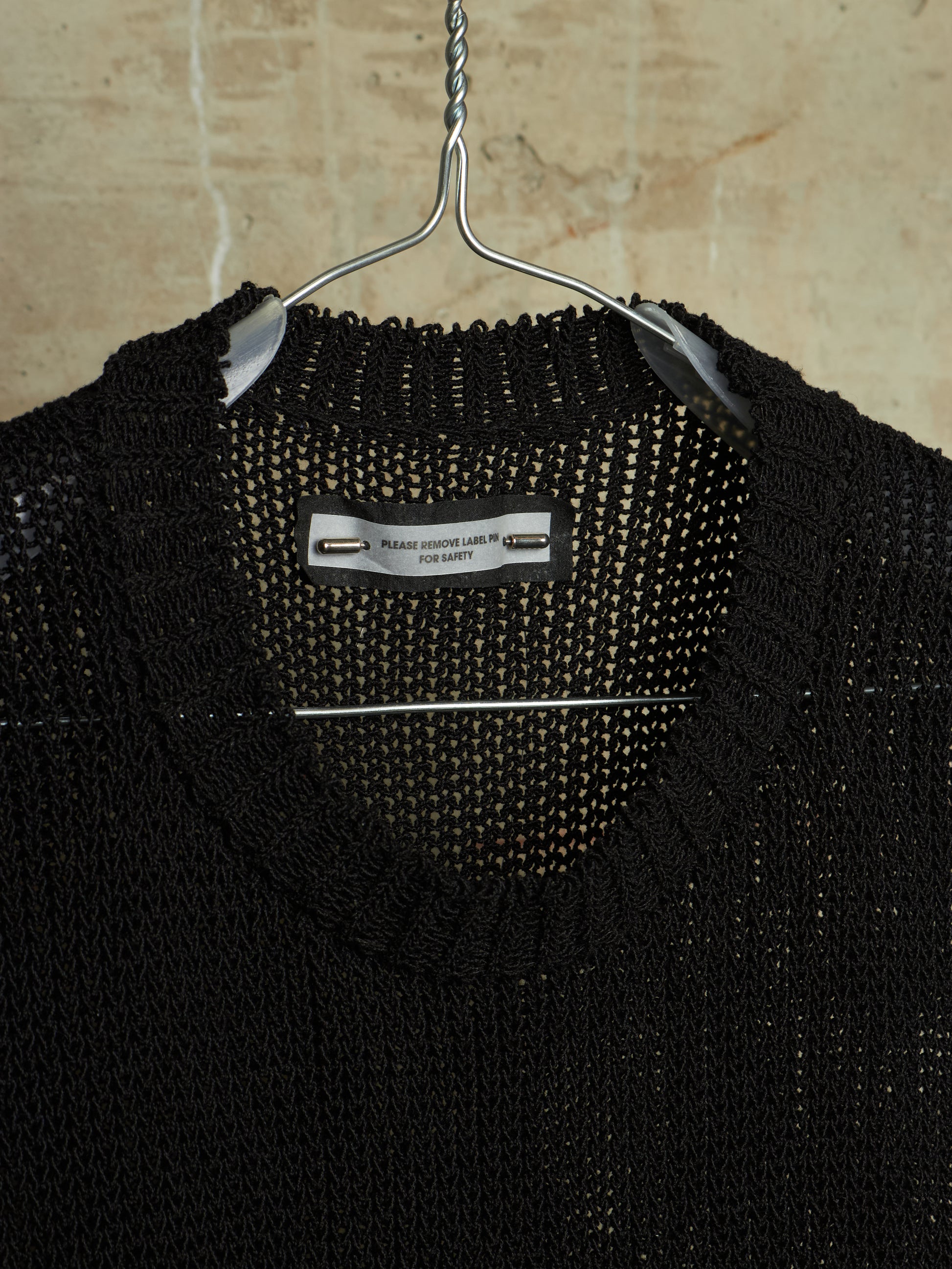A close-up of a black knit sweater hanging on a metal hanger against a beige stone wall. The inner neck label reads 'PERRY FASHION LABEL LTD FOR LATE'.