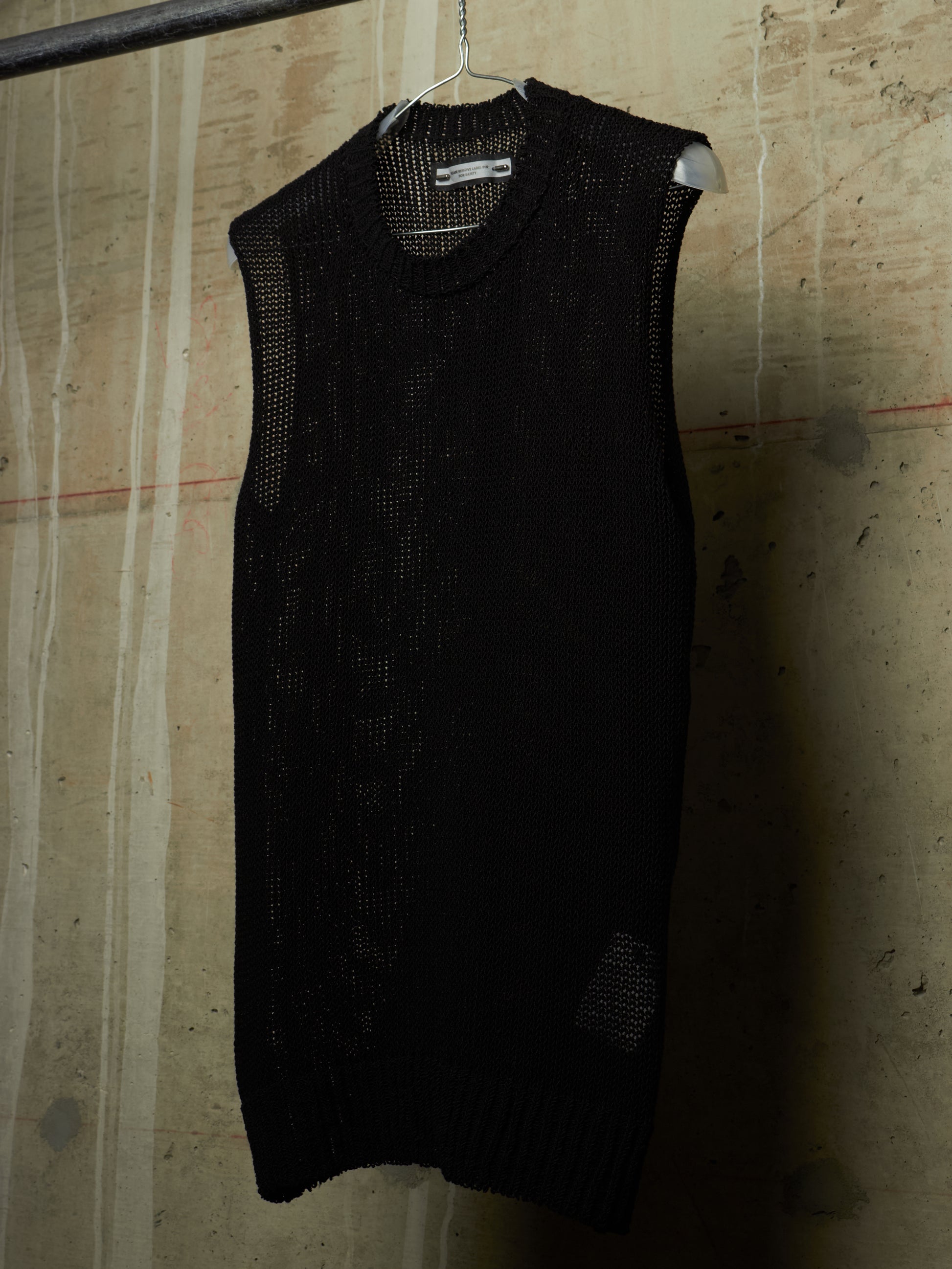 A black sleeveless open-knit sweater vest hanging on a metal hanger against a textured concrete wall.