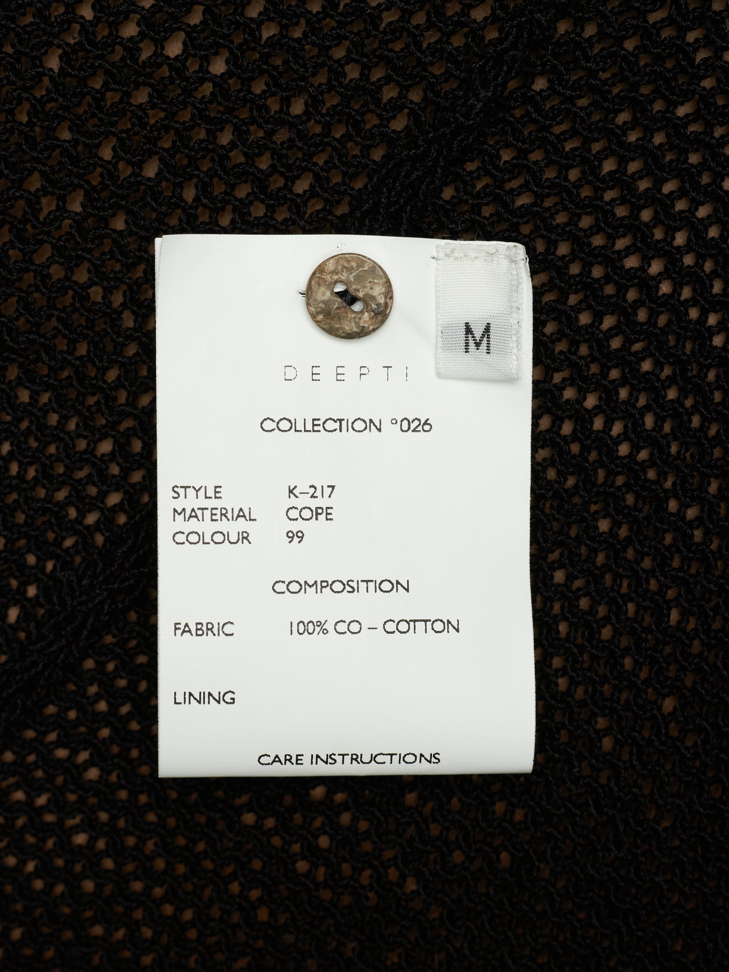 A white fabric care label with printed details and a metal button, placed on a black textured knit fabric background.