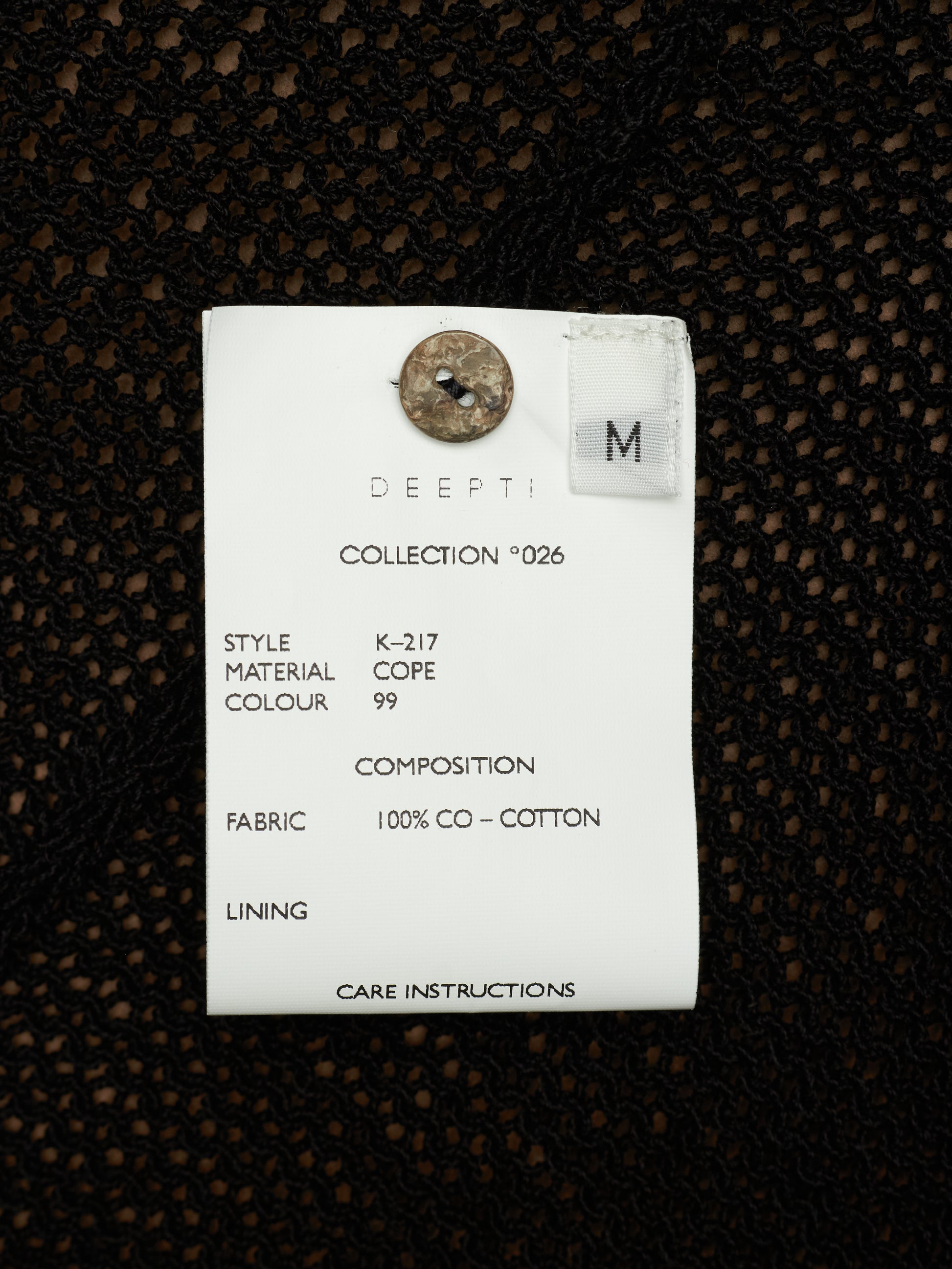 A white fabric care label with printed details and a metal button, placed on a black textured knit fabric background.