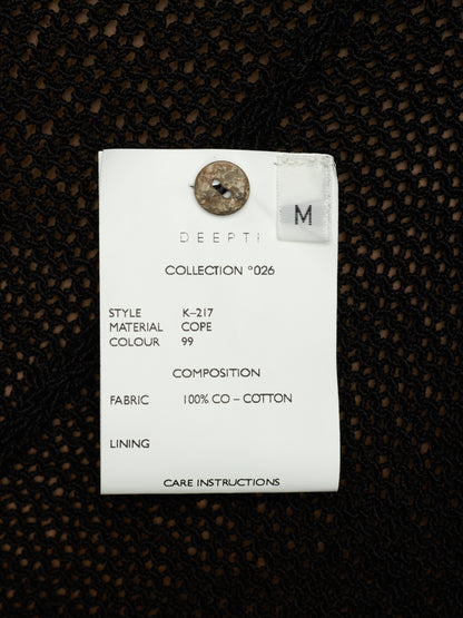 A white fabric care label with printed details and a metal button, placed on a black textured knit fabric background.