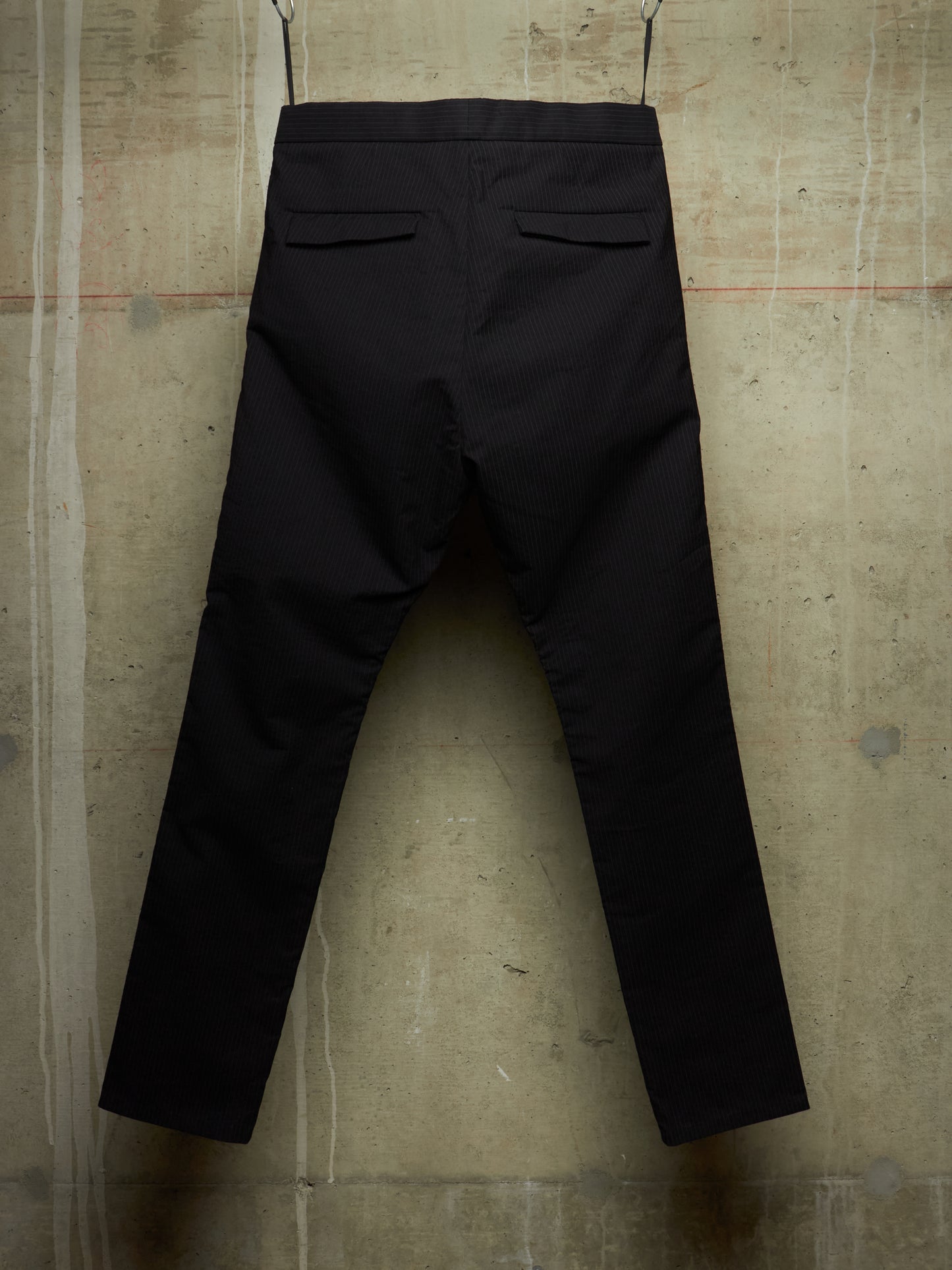 Black trousers shown from the back, hanging against a textured concrete wall.