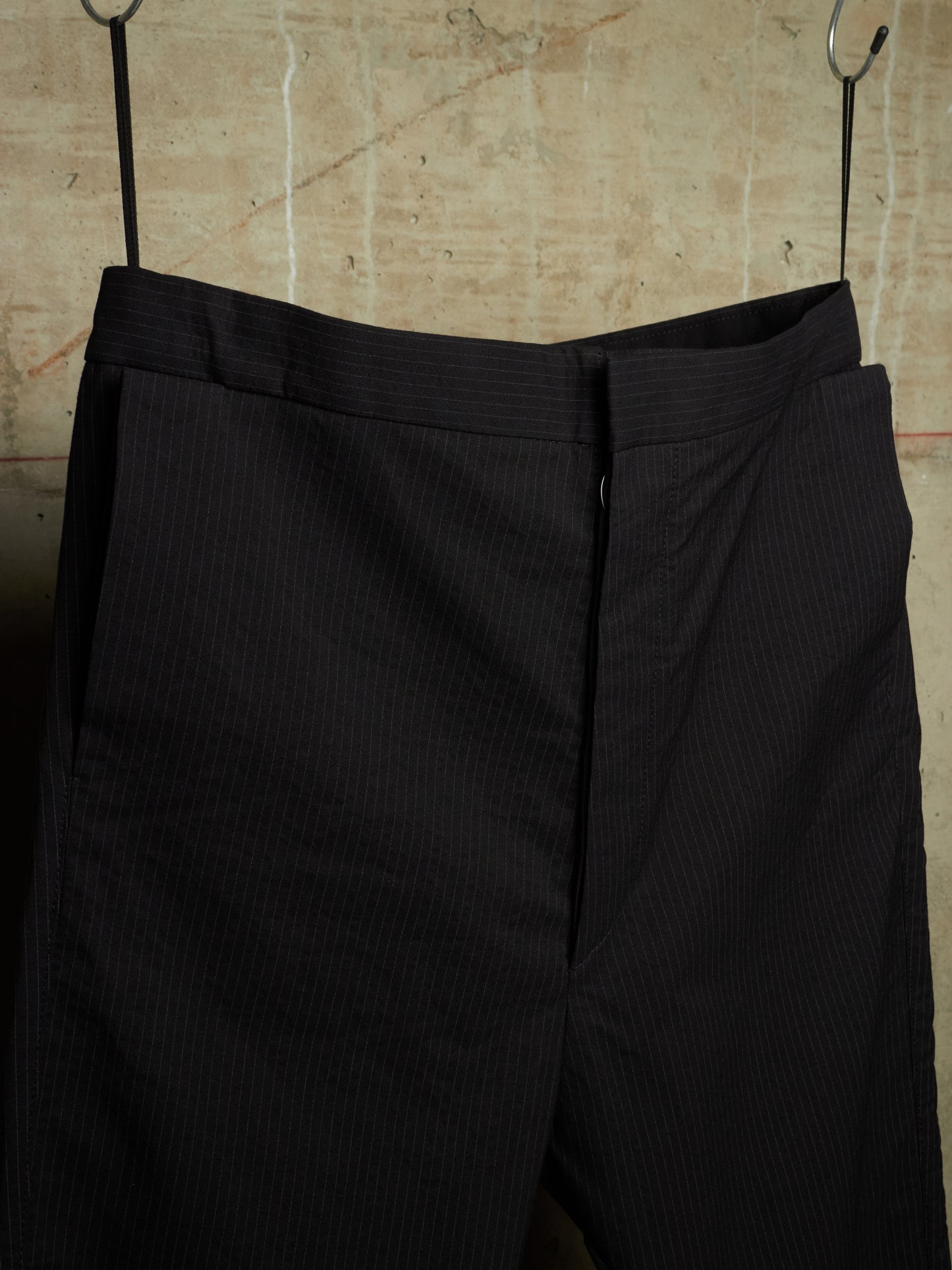 Black pinstriped trousers hanging from metal hooks against a textured beige wall.