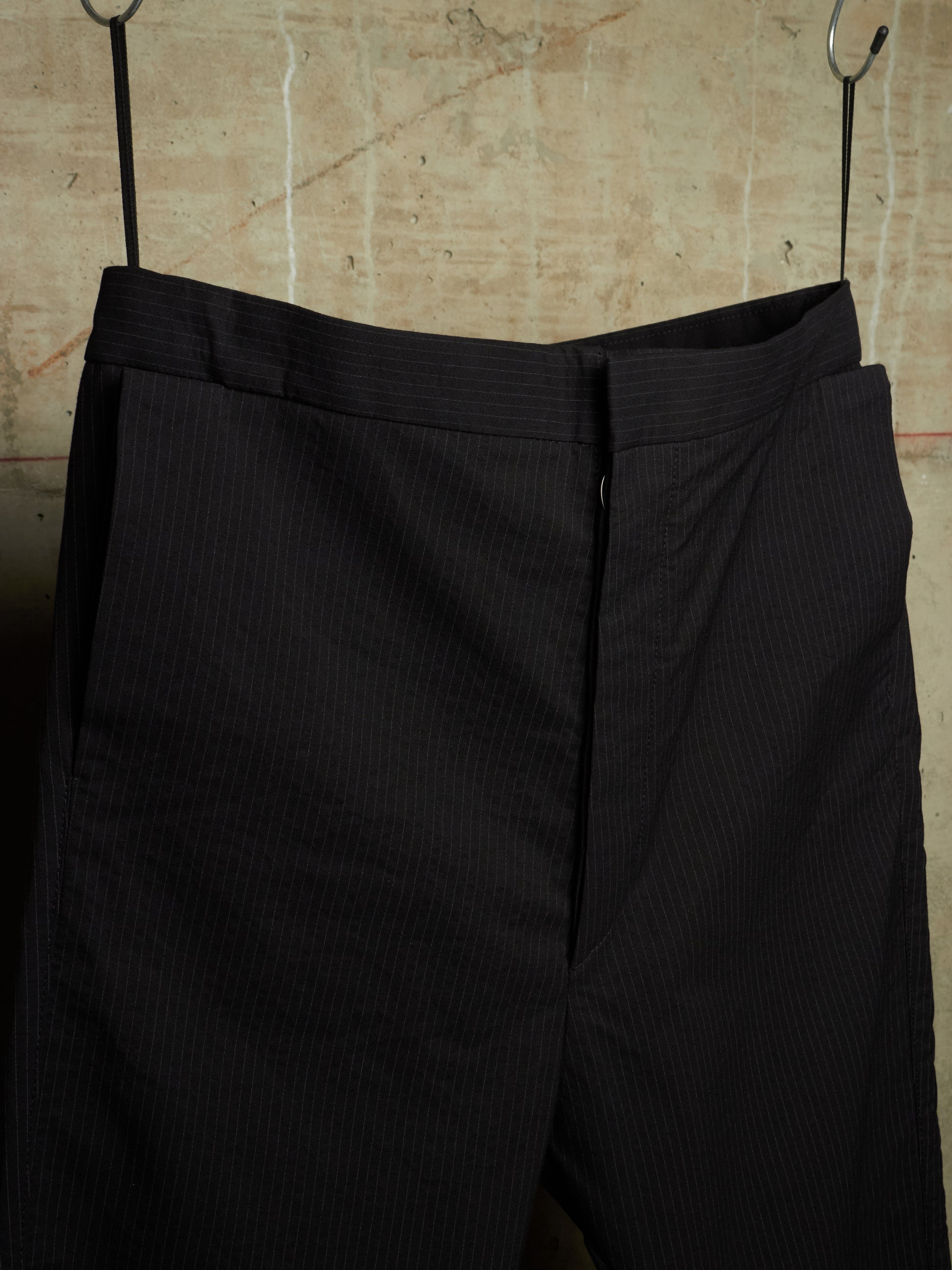Black pinstriped trousers hanging from metal hooks against a textured beige wall.