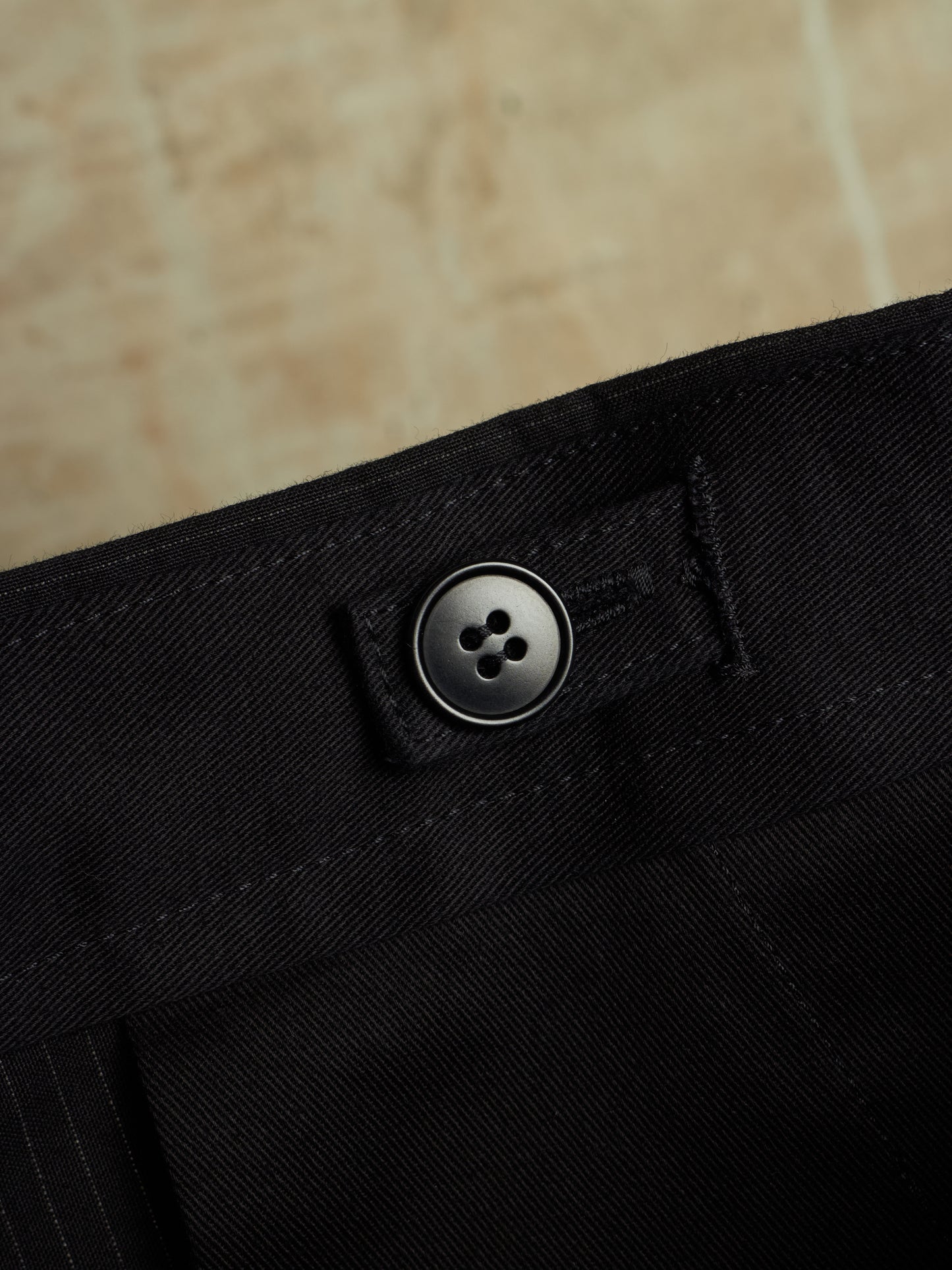 Close-up of black trousers showing the waistband with a single dark button closure and stitched detail, set against a warm beige stone background.