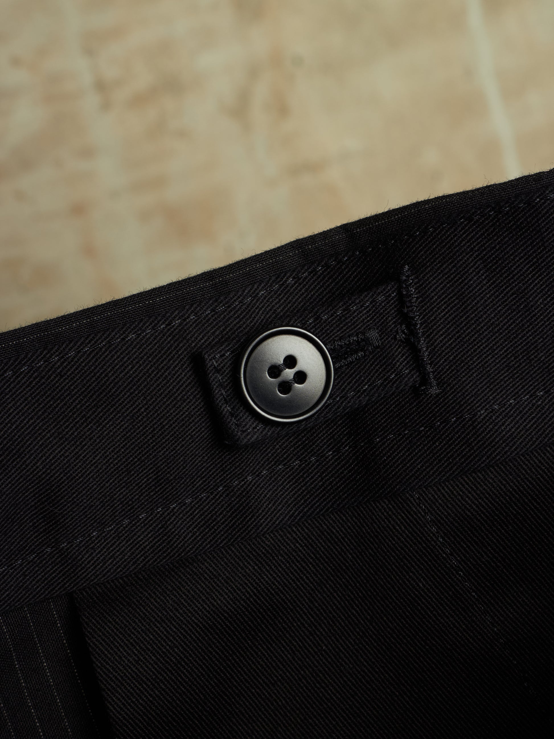 Close-up of black trousers showing the waistband with a single dark button closure and stitched detail, set against a warm beige stone background.