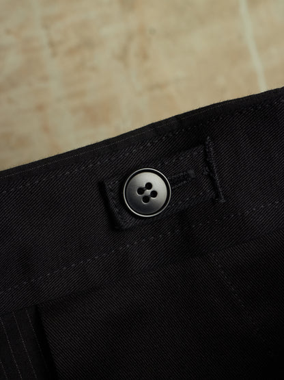 Close-up of black trousers showing the waistband with a single dark button closure and stitched detail, set against a warm beige stone background.