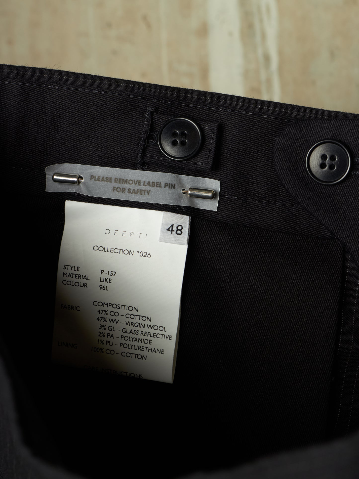 Close-up of the inside waistband of black trousers showing a white brand label for DEEPTI, with black buttons and a metal safety pin attached.