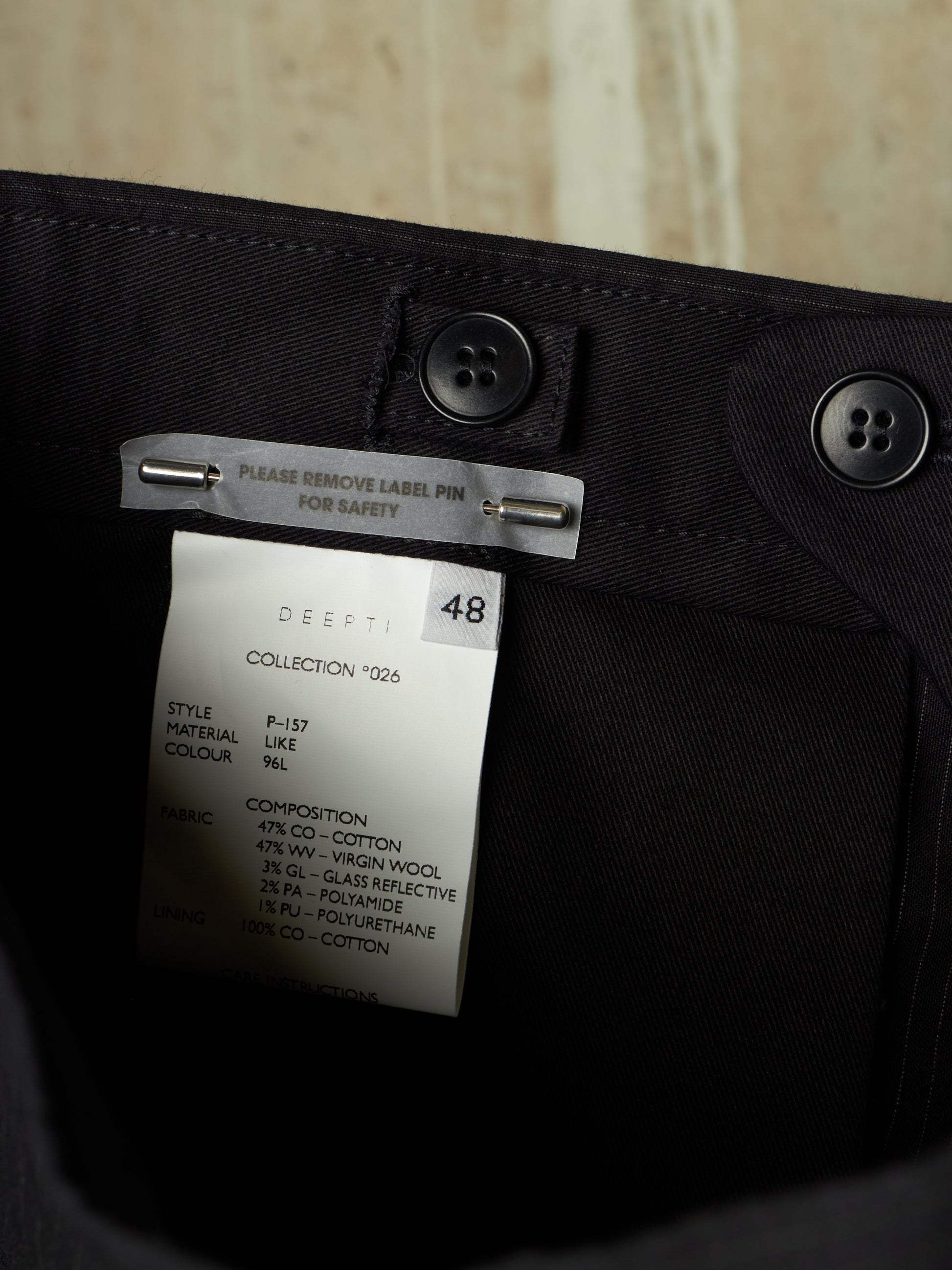 Close-up of the inside waistband of black trousers showing a white brand label for DEEPTI, with black buttons and a metal safety pin attached.