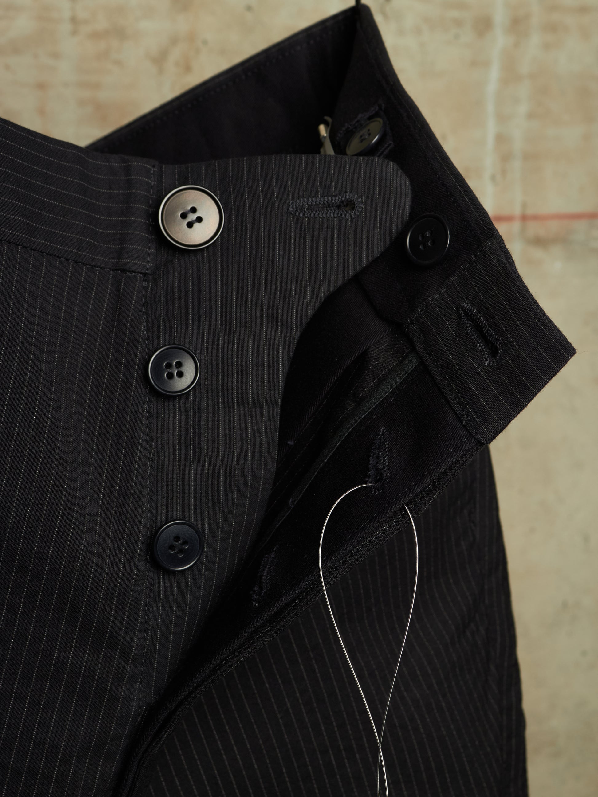 Close-up of a black pinstriped shirt jacket showing the collar, button closure, and thread against a textured beige background.