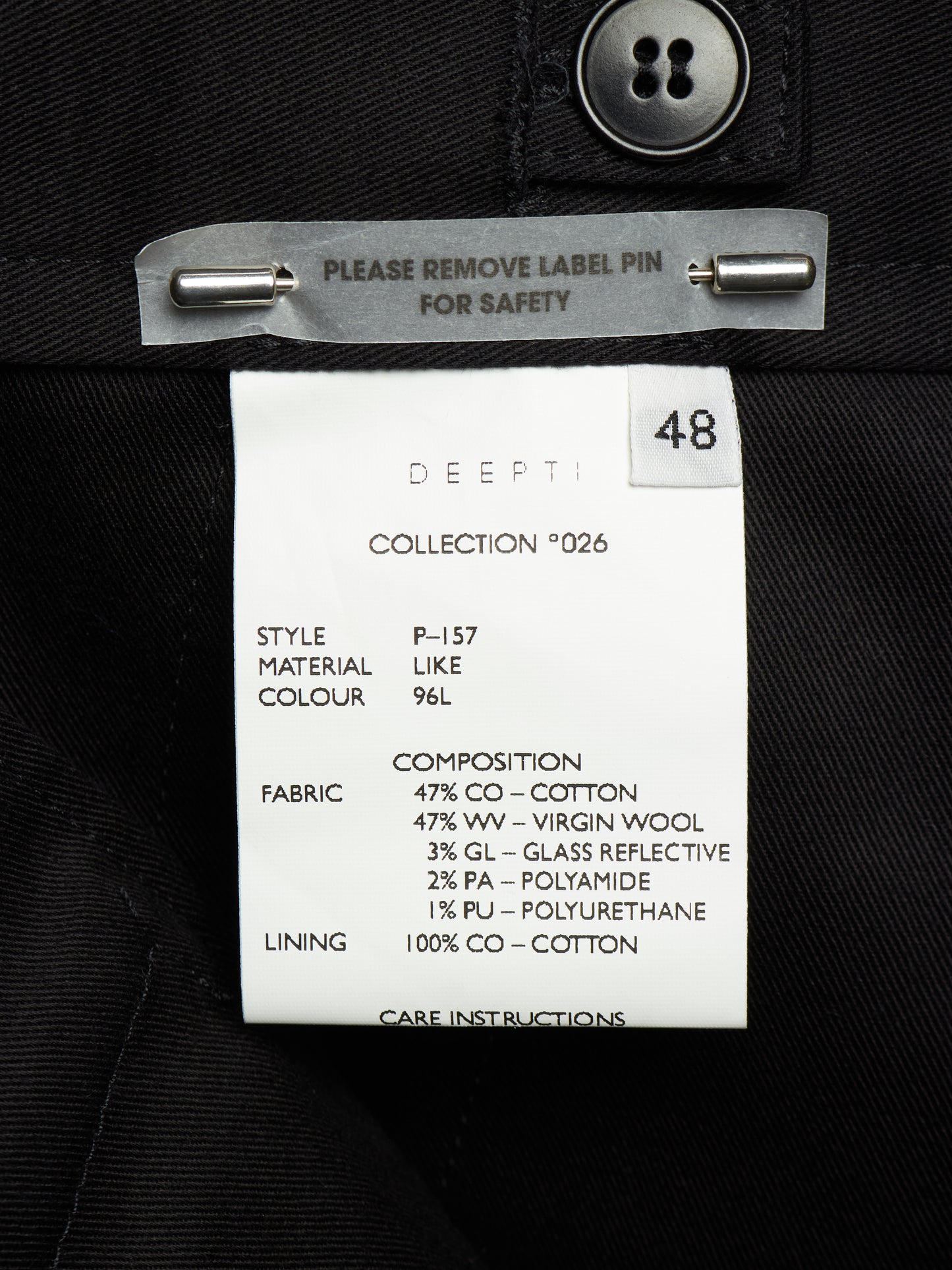 A close-up of a garment's inner neck label reading 'DEEPTI COLLECTION *026' with size 48, attached to a black fabric garment with a metal pin clip.