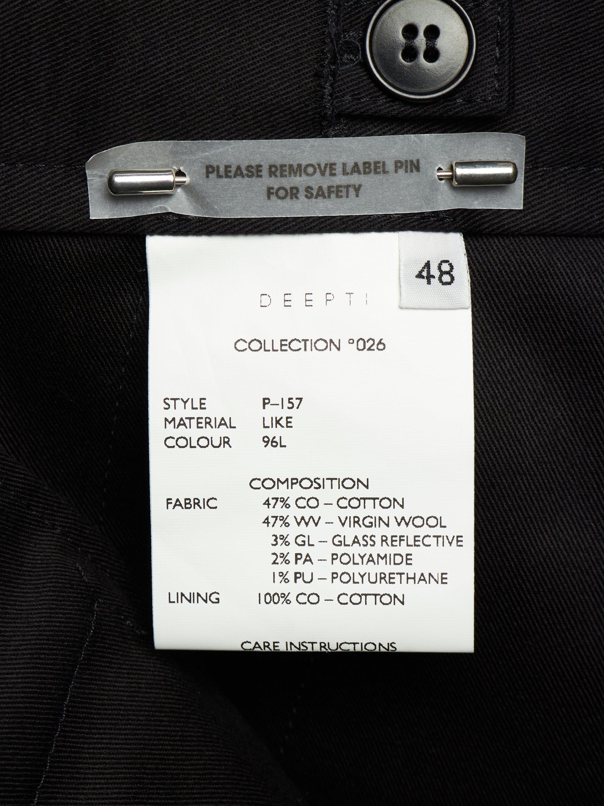 A close-up of a garment's inner neck label reading 'DEEPTI COLLECTION *026' with size 48, attached to a black fabric garment with a metal pin clip.
