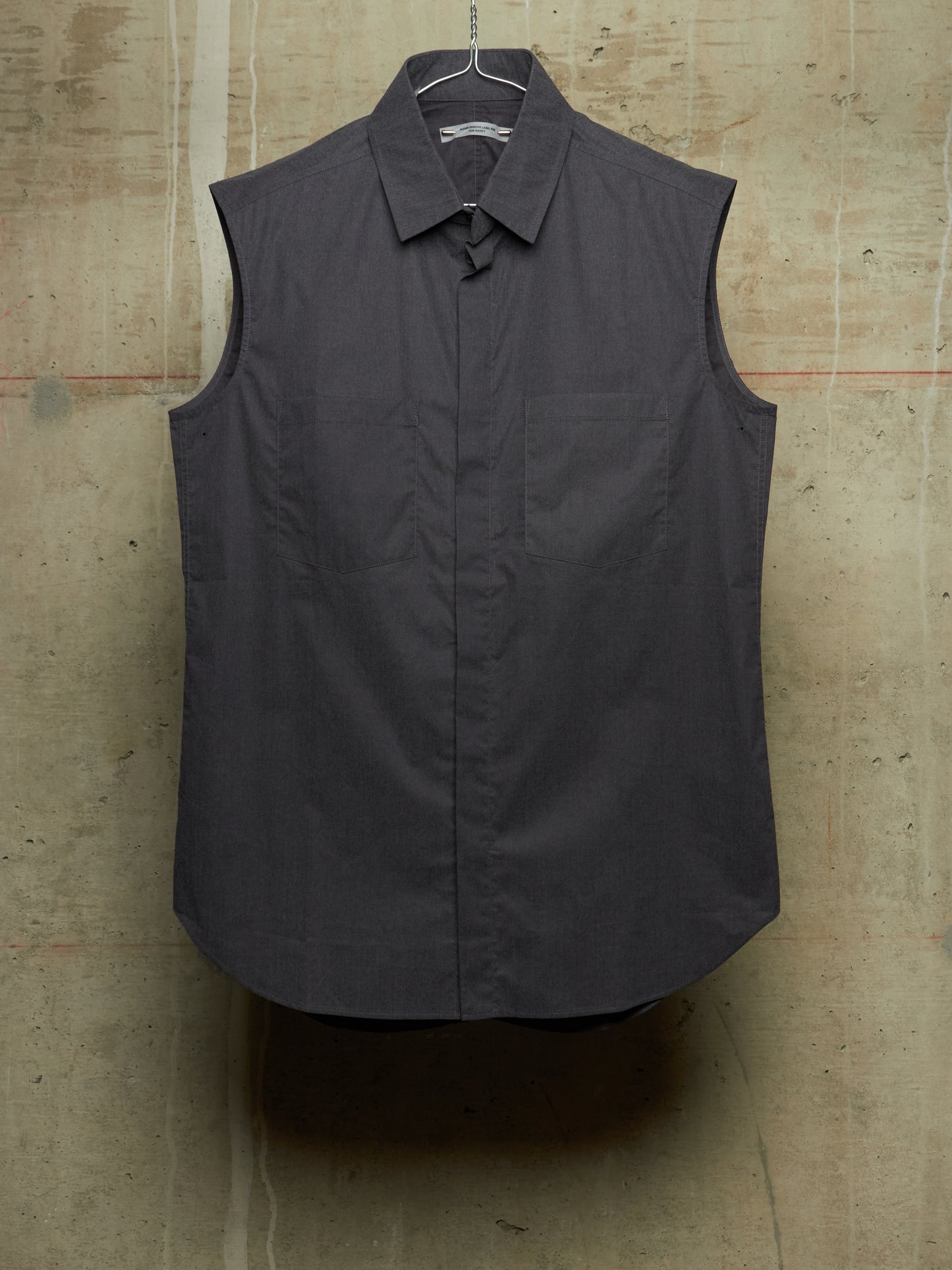 A dark charcoal gray sleeveless button-up shirt with a pointed collar and chest pocket, hanging on a wire hanger against a textured concrete wall.