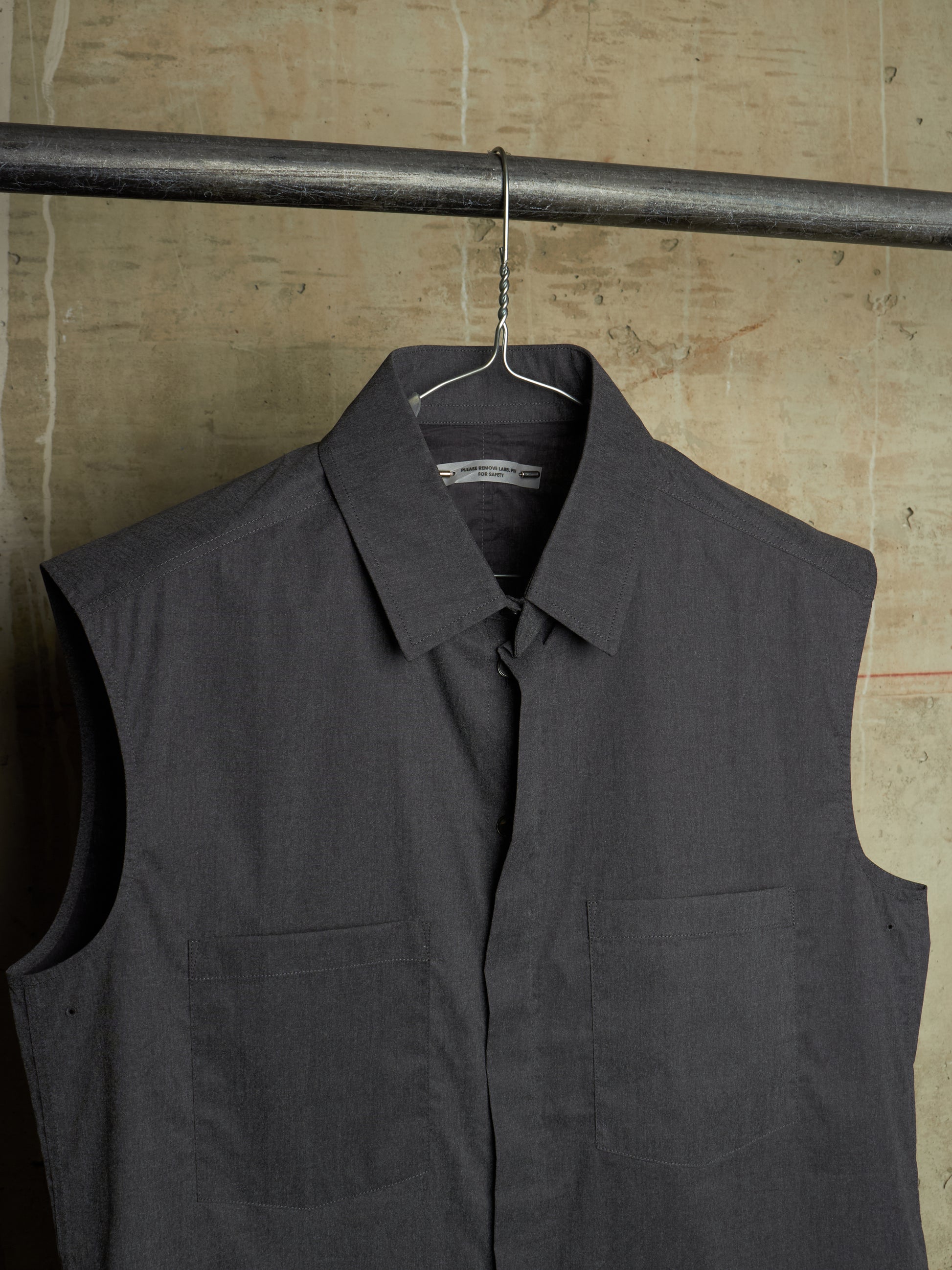 A dark gray sleeveless button-up shirt vest with a pointed collar and two front chest pockets, hanging on a metal hanger from a horizontal clothing rack against a textured beige wall.