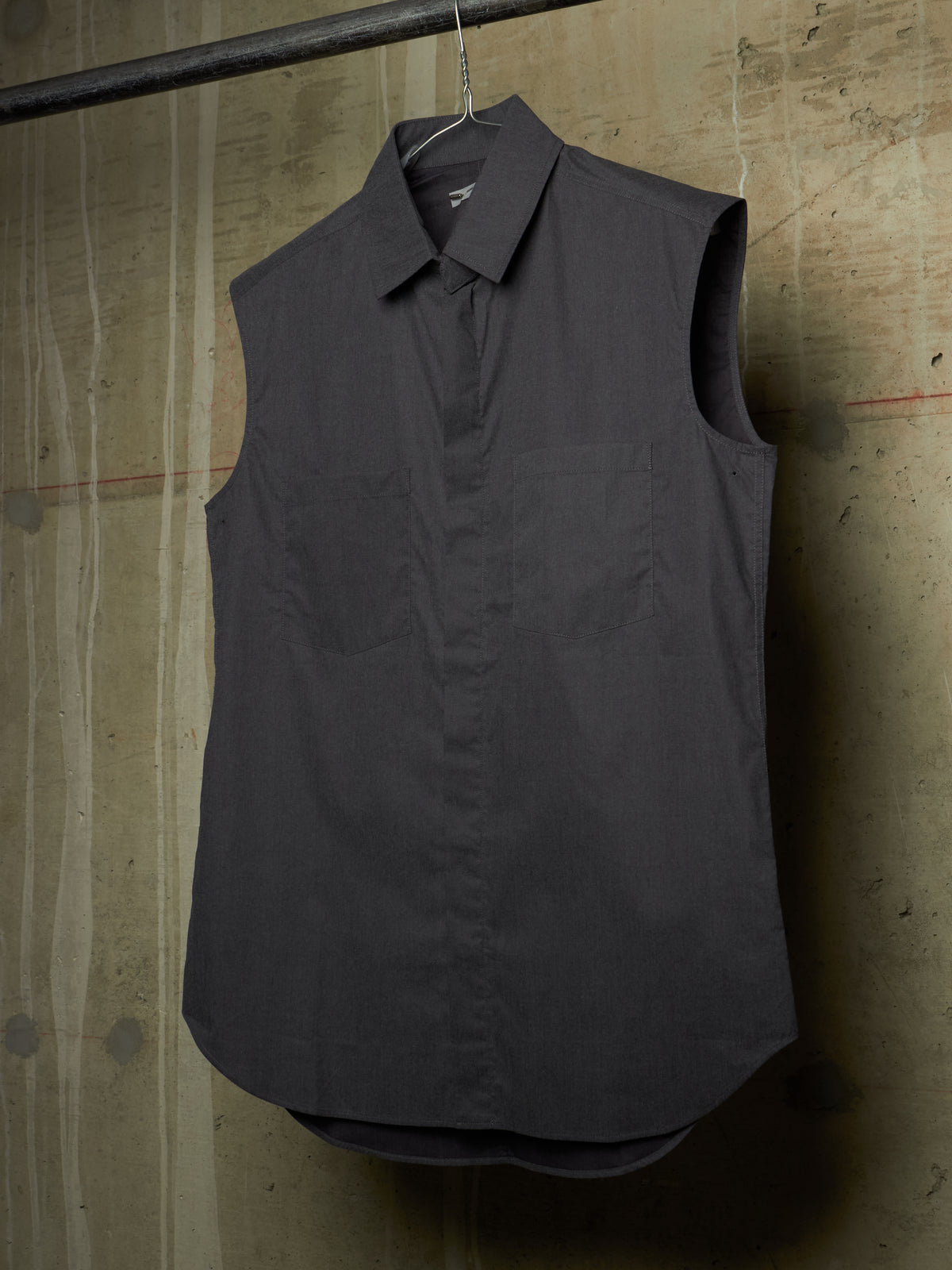 A dark charcoal gray sleeveless collared shirt with a button-down front and chest pocket, hanging on a metal hanger from a black clothing rack against a textured concrete wall.