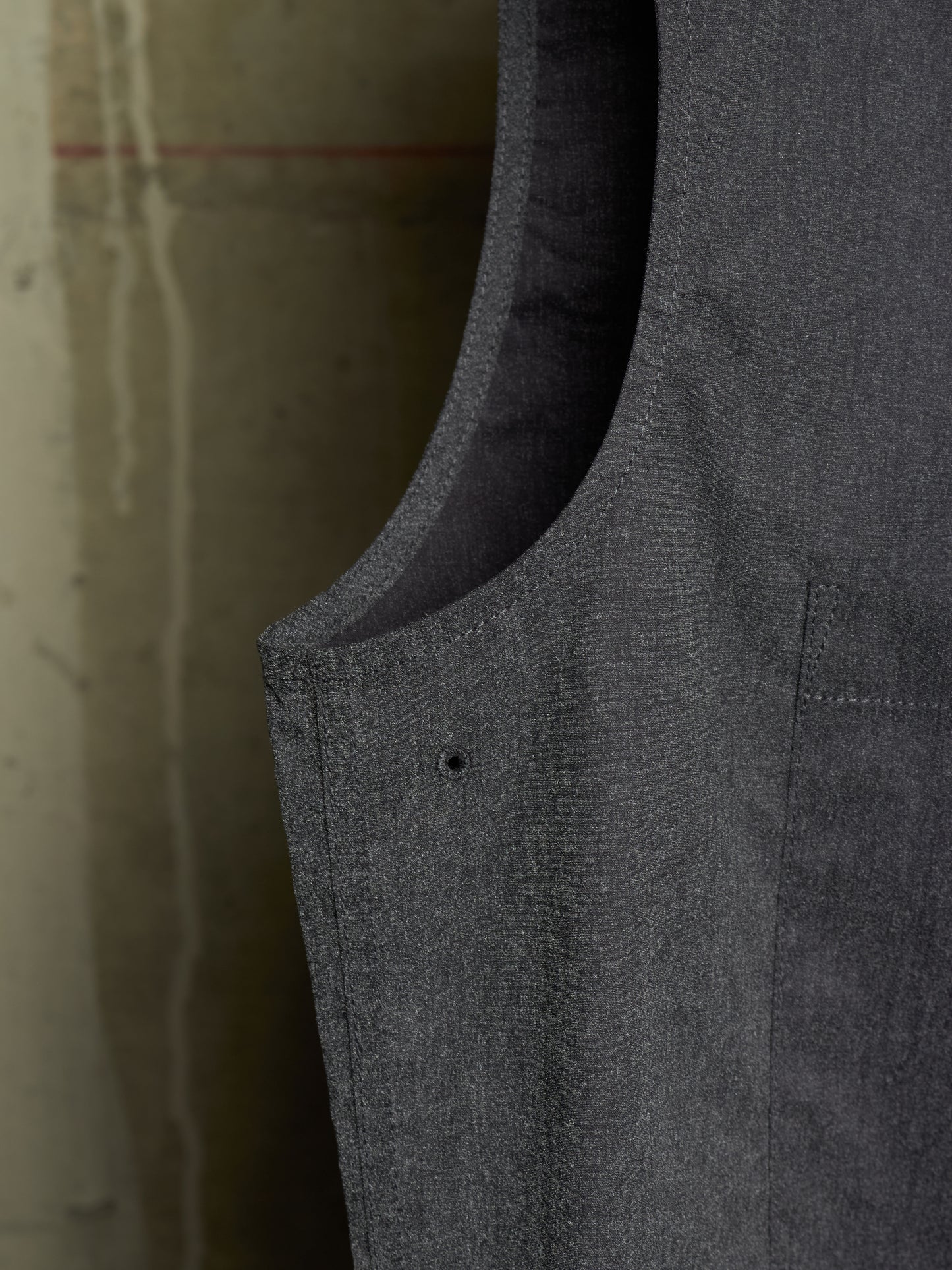 Close-up detail shot of a dark gray textured fabric garment, showing the inner lining, curved neckline, and a small button detail against a neutral concrete background.