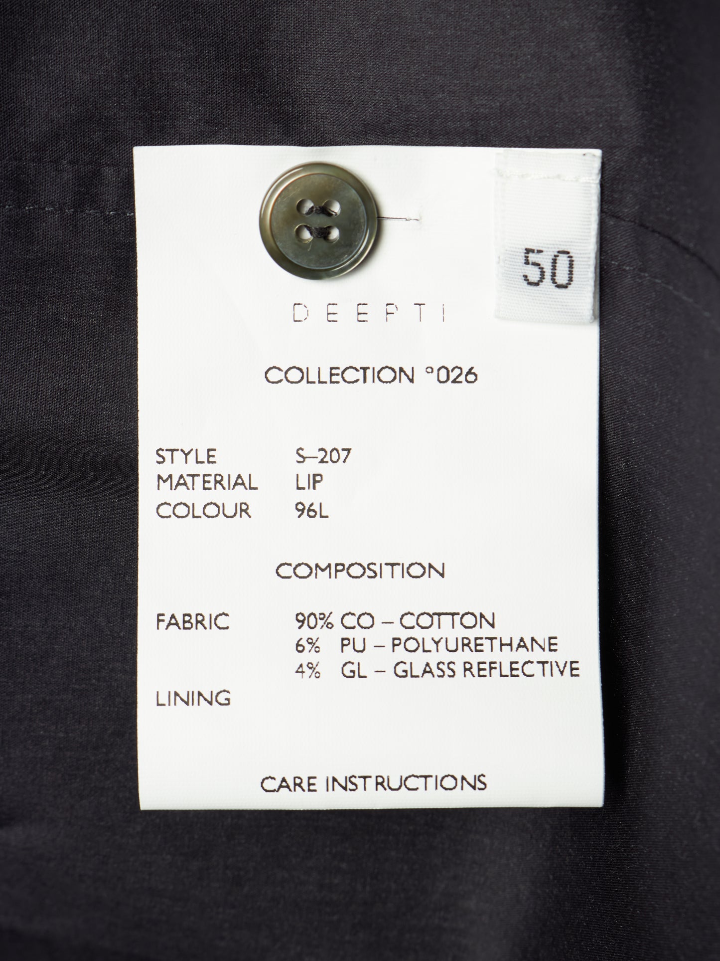 A white fabric care label with black text attached to a dark navy or black garment, featuring a metal button and a small white sticker marked 50.