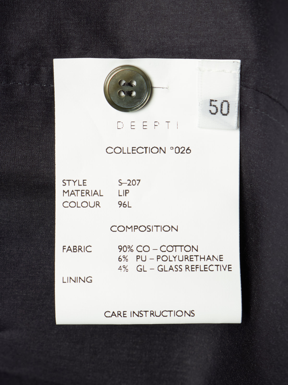 A white fabric care label with black text attached to a dark navy or black garment, featuring a metal button and a small white sticker marked 50.