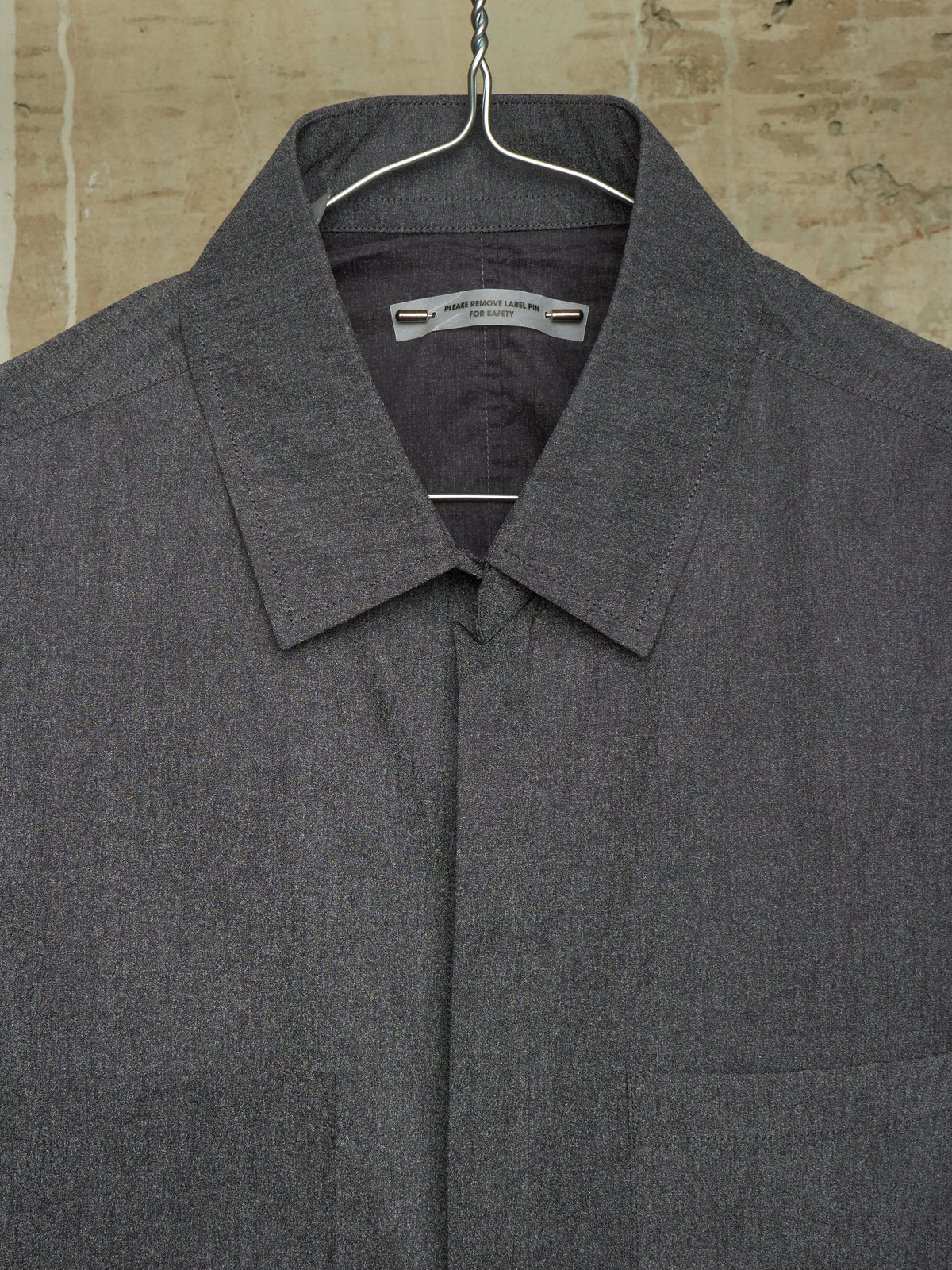 A dark gray textured collared shirt jacket hanging on a metal hanger against a rustic beige stone wall background.