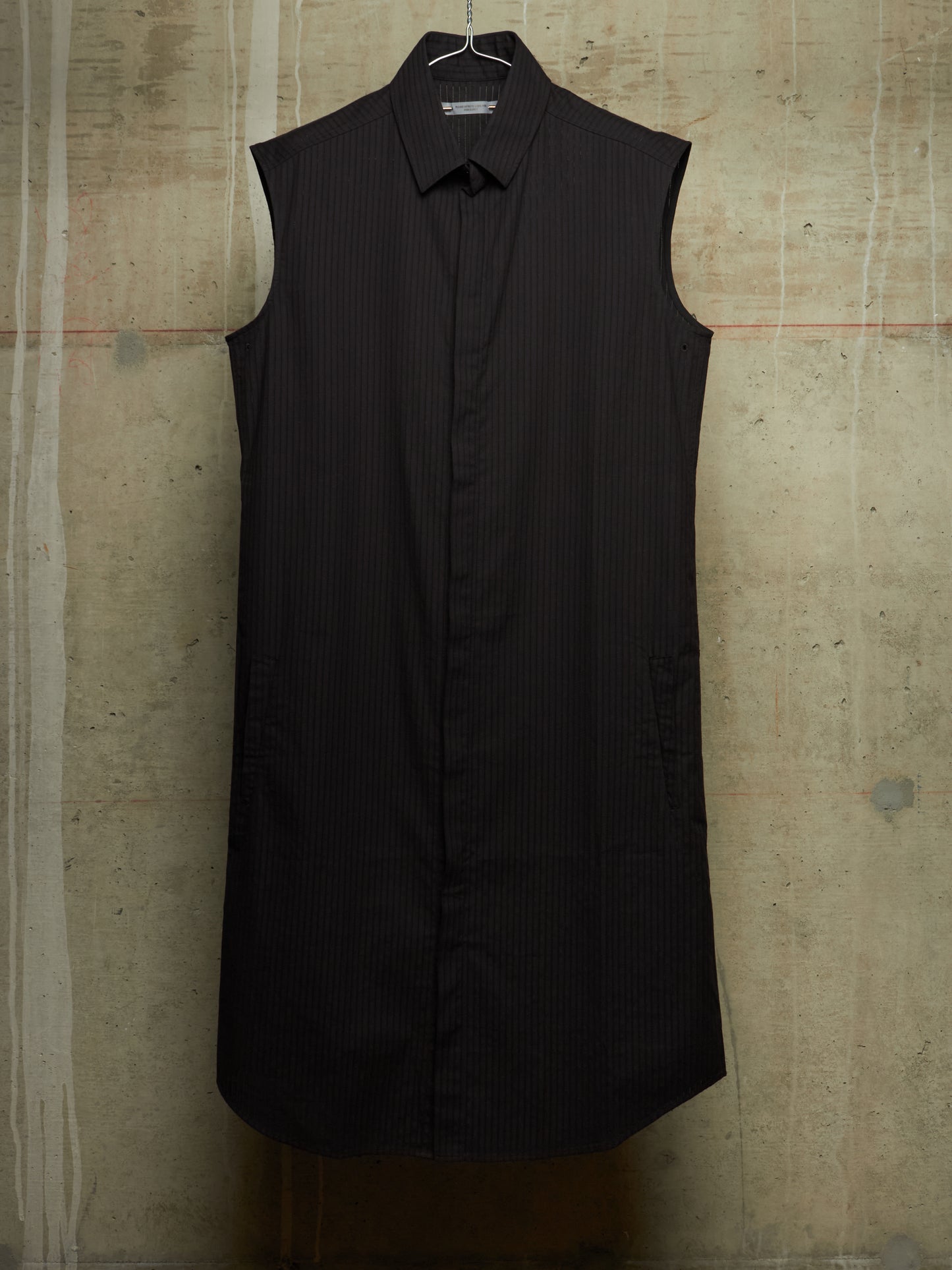 A sleeveless black pinstriped button-down shirt dress with a pointed collar, hanging on a wire hanger against a textured concrete wall.