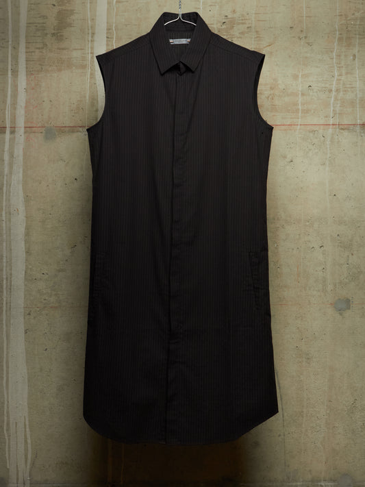 A sleeveless black pinstriped button-down shirt dress with a pointed collar, hanging on a wire hanger against a textured concrete wall.
