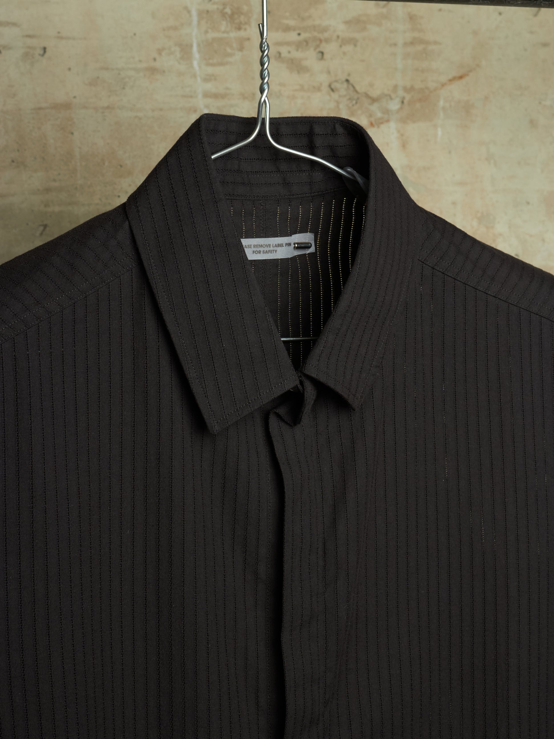 A dark brown pinstriped shirt hanging on a metal hanger against a textured beige stone wall.