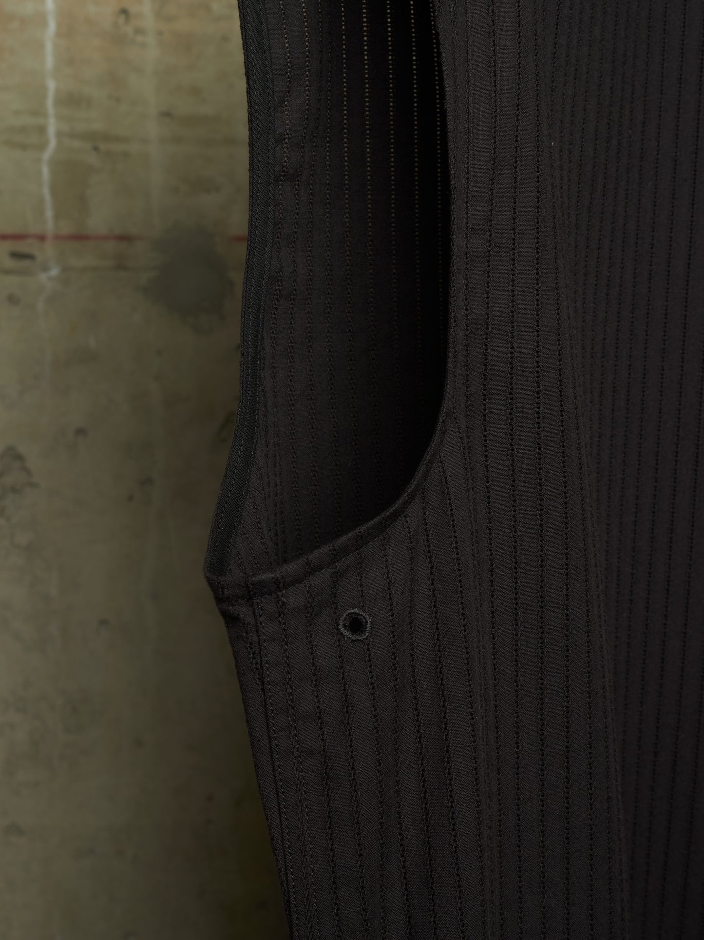 Close-up detail of a dark charcoal gray pinstriped garment, showing the texture of the fabric, a side pocket seam, and a metal button, set against a concrete wall background.
