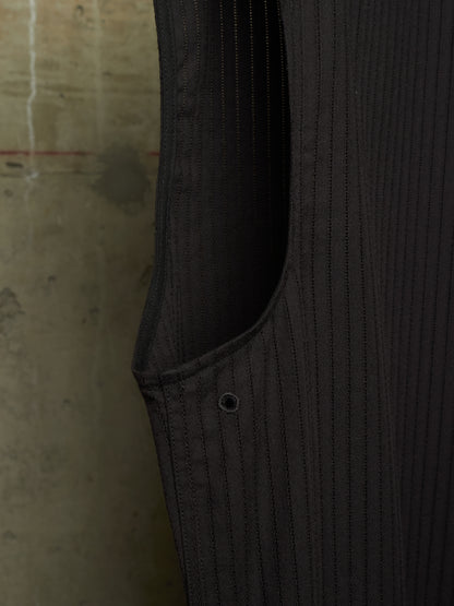 Close-up detail of a dark charcoal gray pinstriped garment, showing the texture of the fabric, a side pocket seam, and a metal button, set against a concrete wall background.