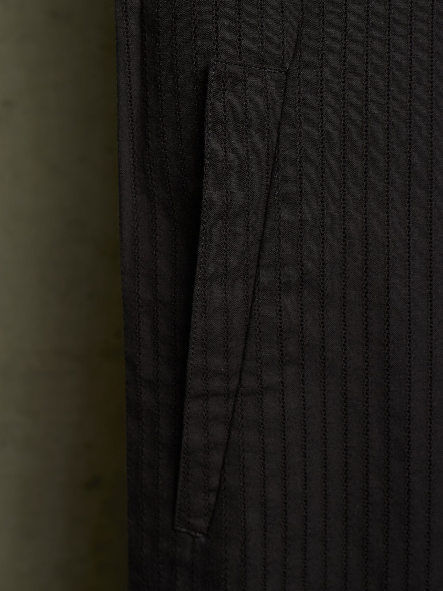 Close-up of a dark charcoal gray pinstriped fabric garment, showing a vertical pocket seam and fine stitched texture.