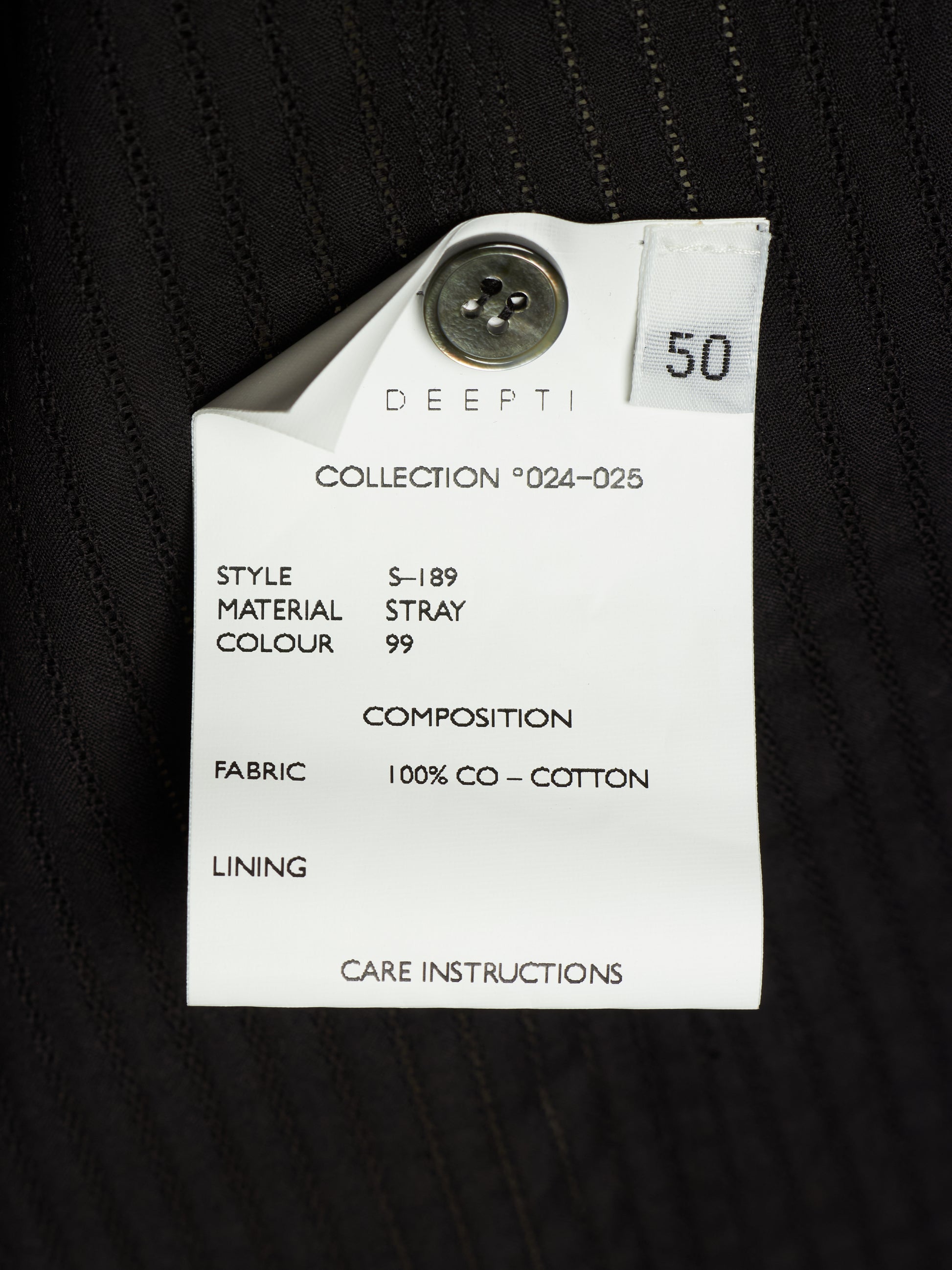 A close-up of a white paper garment tag attached to a black pinstriped fabric garment. The tag is printed with details including 'DEEPTI', 'COLLECTION *024-025', 'STYLE S-189', 'MATERIAL STRAY', 'COLOUR 99', 'FABRIC 100% CO - COTTON', and 'CARE INSTRUCTIONS'.