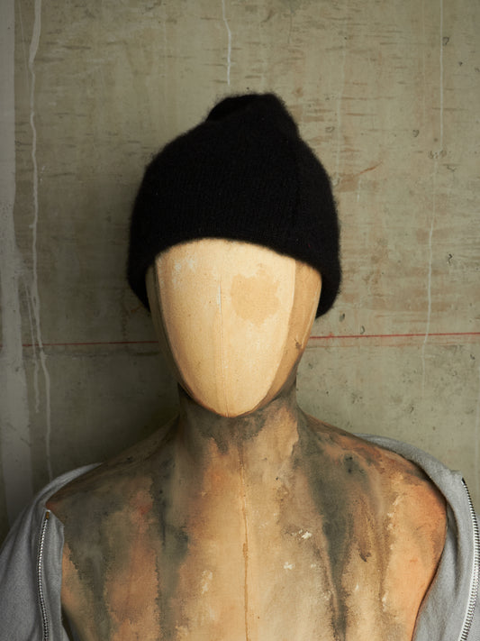 A black knit beanie hat displayed on a wooden mannequin torso against a textured concrete wall.