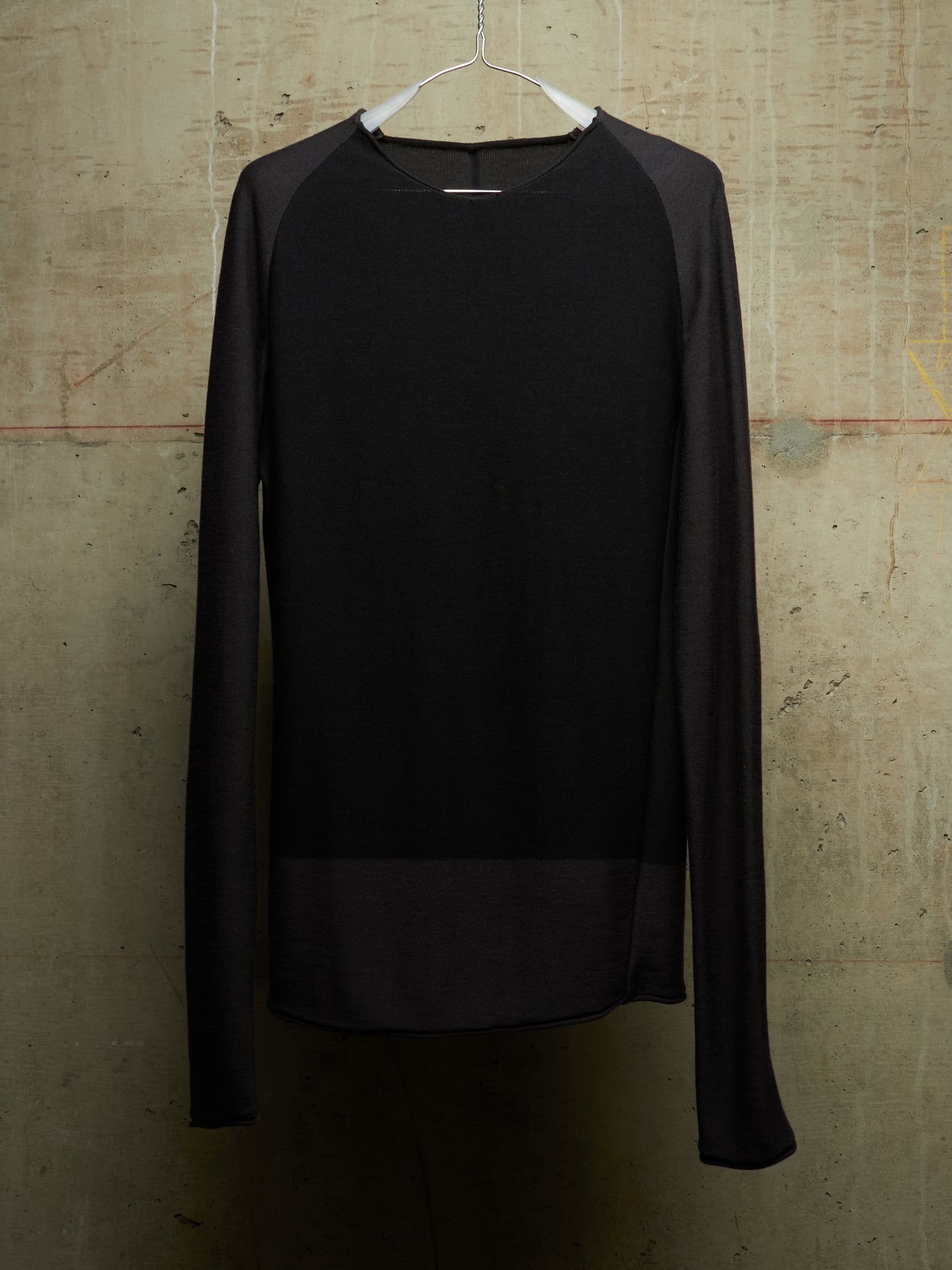 A black long-sleeve knit top with sheer mesh raglan sleeves and a sheer mesh hem panel, hanging on a white hanger against a textured concrete wall.