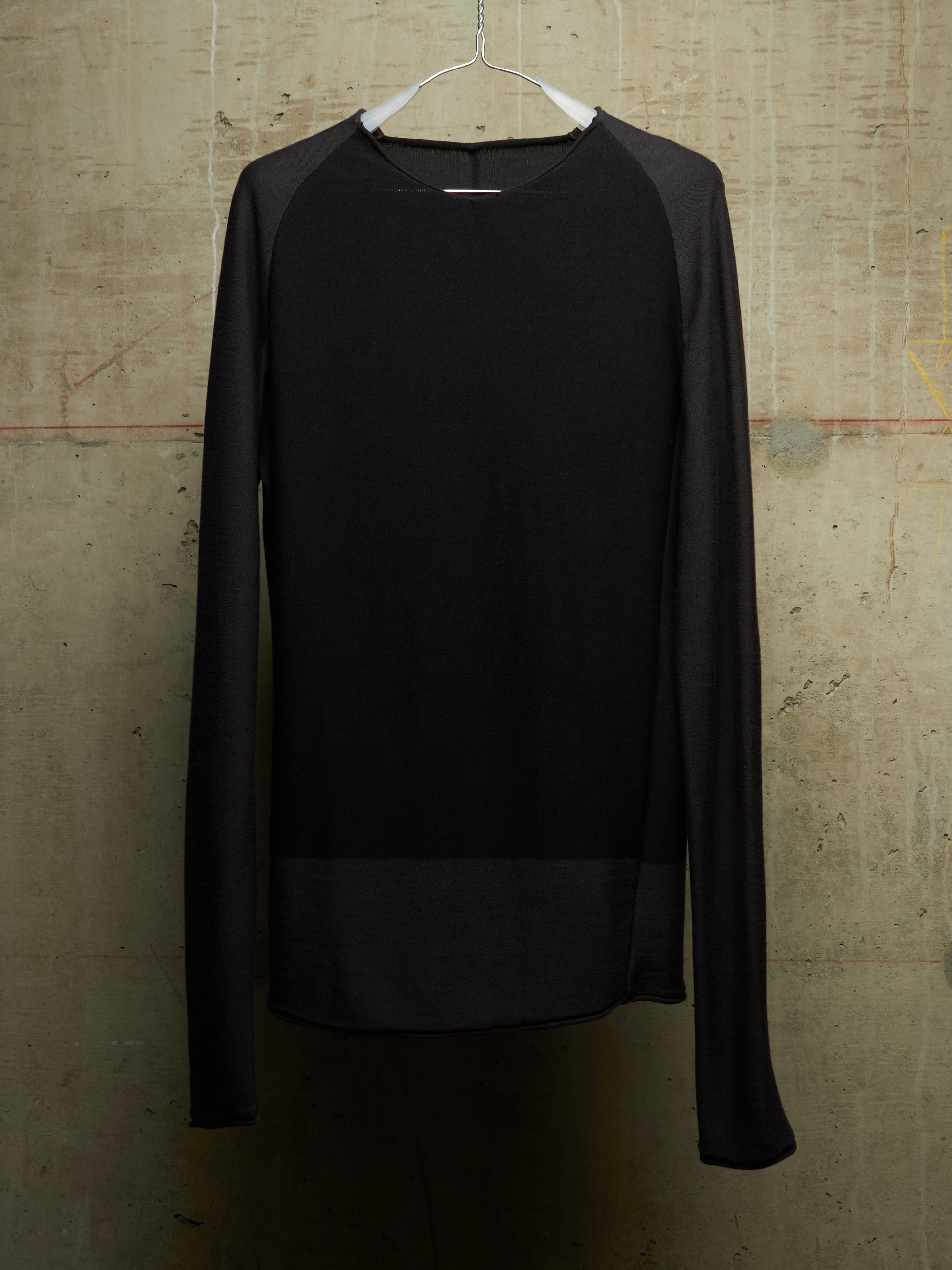 A black long-sleeve knit top with sheer mesh raglan sleeves and a sheer mesh hem panel, hanging on a white hanger against a textured concrete wall.