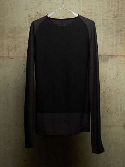 A black long-sleeve knit top with sheer mesh raglan sleeves and a sheer mesh hem panel, hanging on a white hanger against a textured concrete wall.