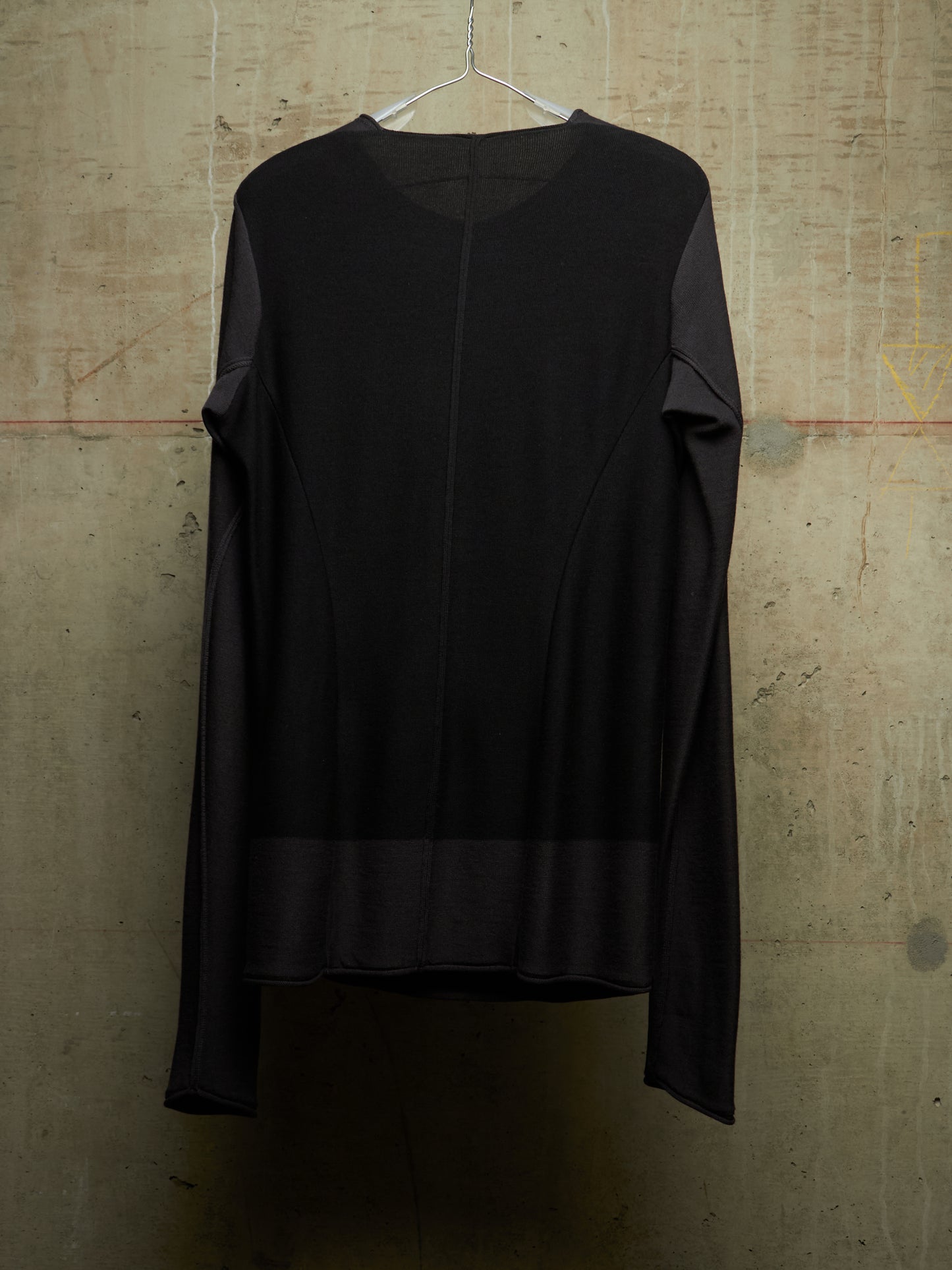 A black long-sleeve top with a crew neck and asymmetrical draped sleeve detail, hanging on a wire hanger against a textured concrete wall.