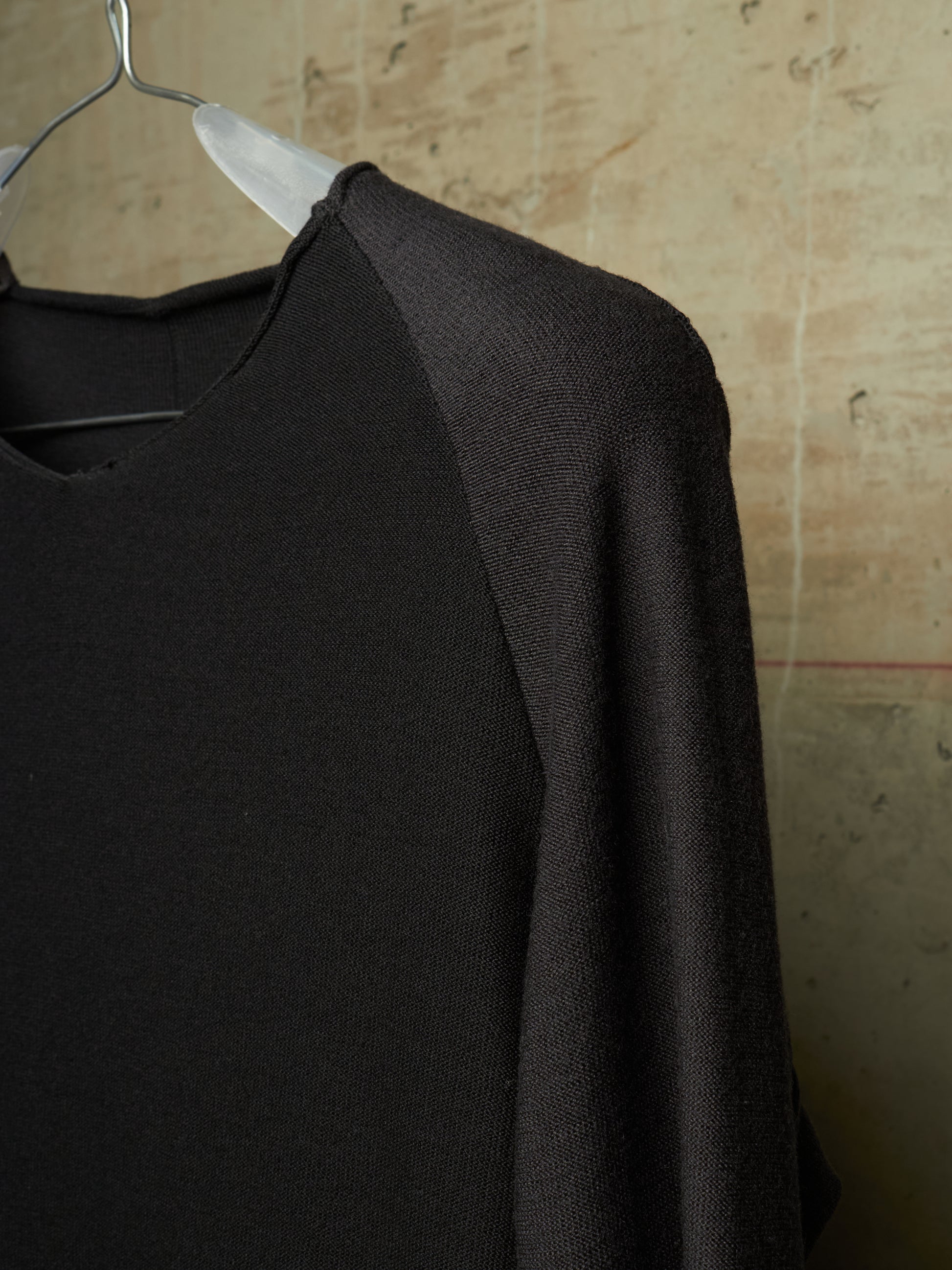 A close-up of a black knit raglan sweater hanging on a clear plastic hanger against a textured beige wall.