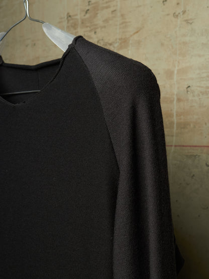 A close-up of a black knit raglan sweater hanging on a clear plastic hanger against a textured beige wall.