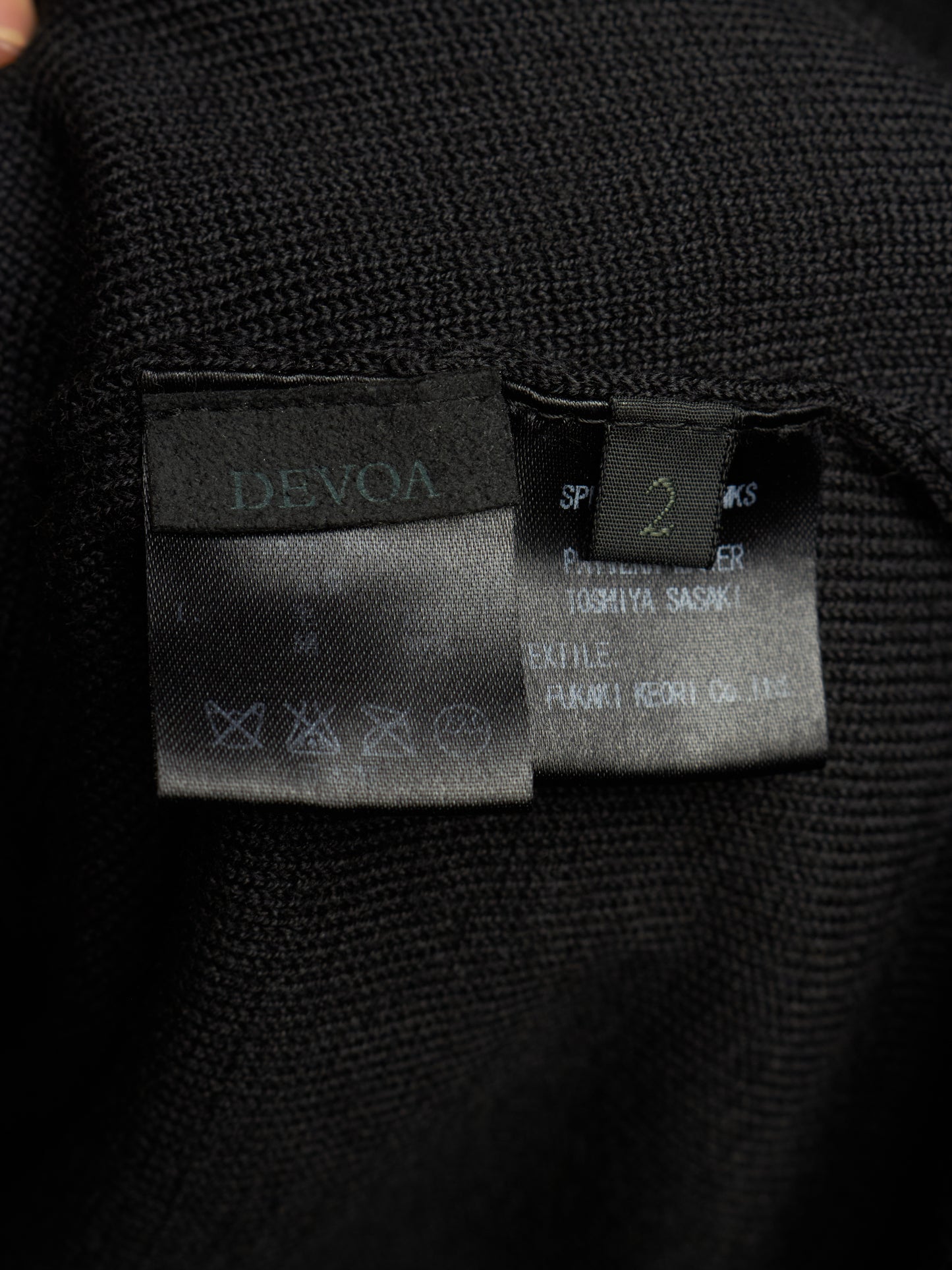 Close-up of a black knit garment showing two interior labels: a larger Devoa label on the left and a smaller size/manufacturer label on the right.