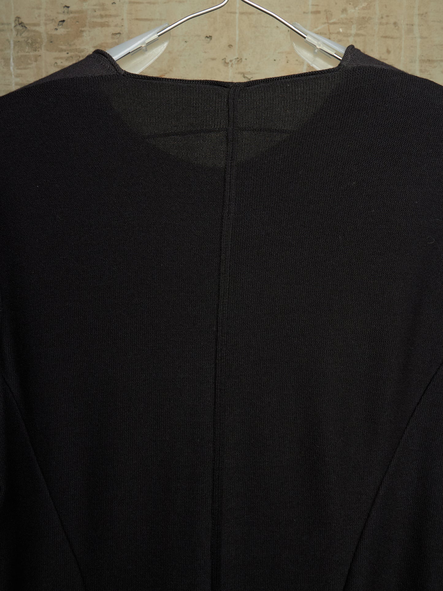 A close-up of a black knit top hanging on a clear plastic hanger against a light beige textured wall.