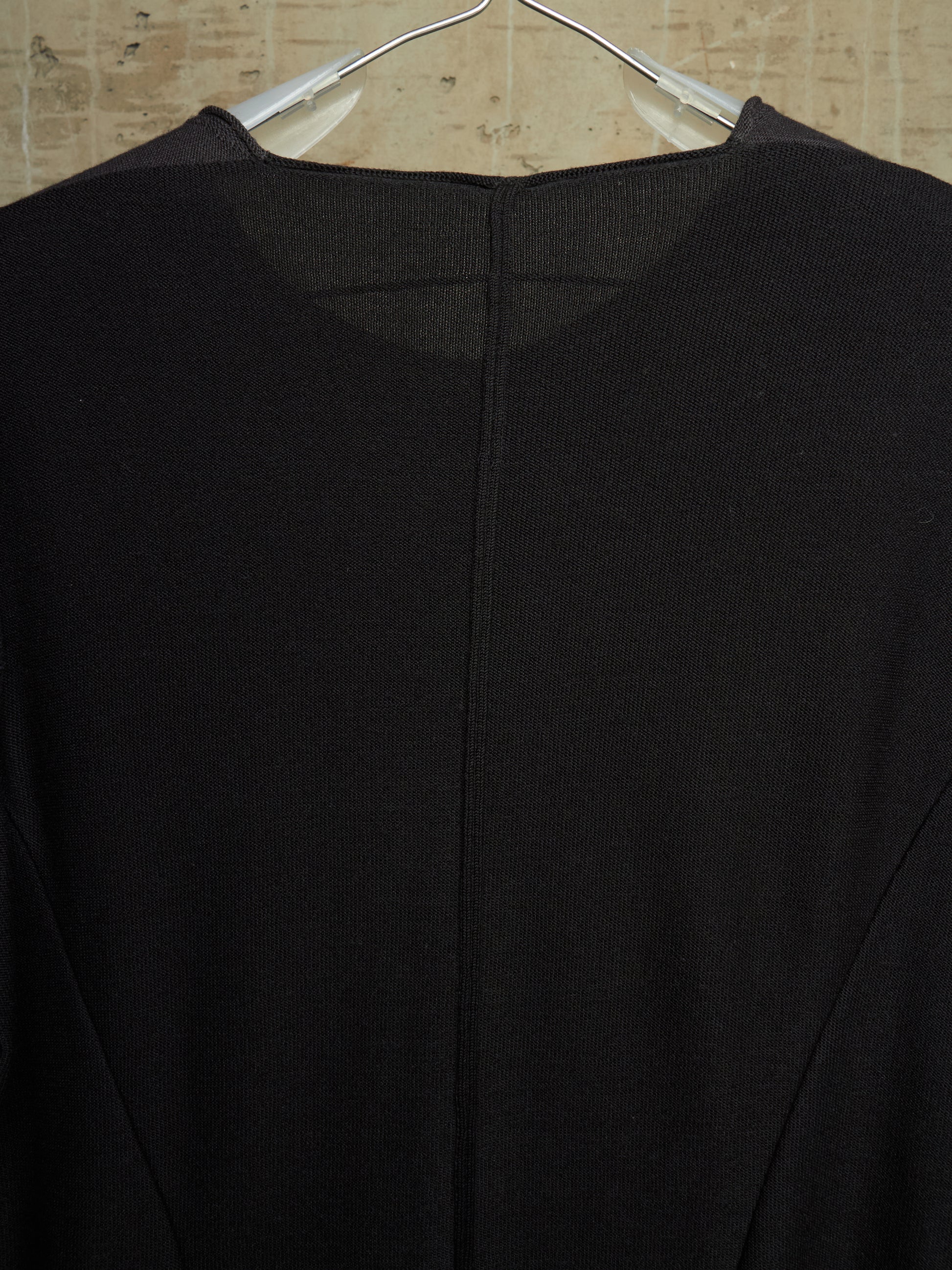 A close-up of a black knit top hanging on a clear plastic hanger against a light beige textured wall.